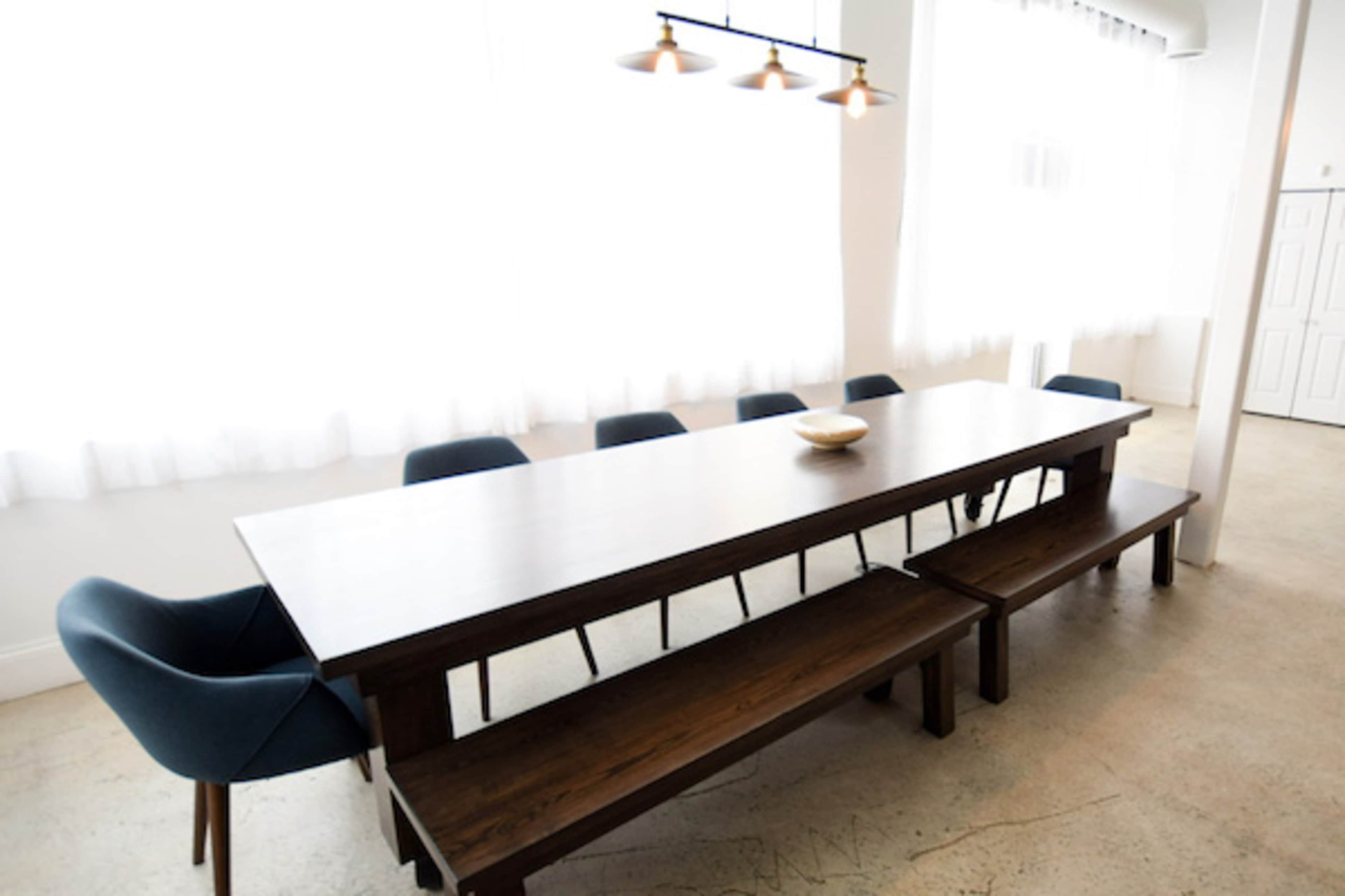A long wooden dining table with a central bowl is surrounded by blue upholstered chairs and a wooden bench in a bright room with sheer white curtains.