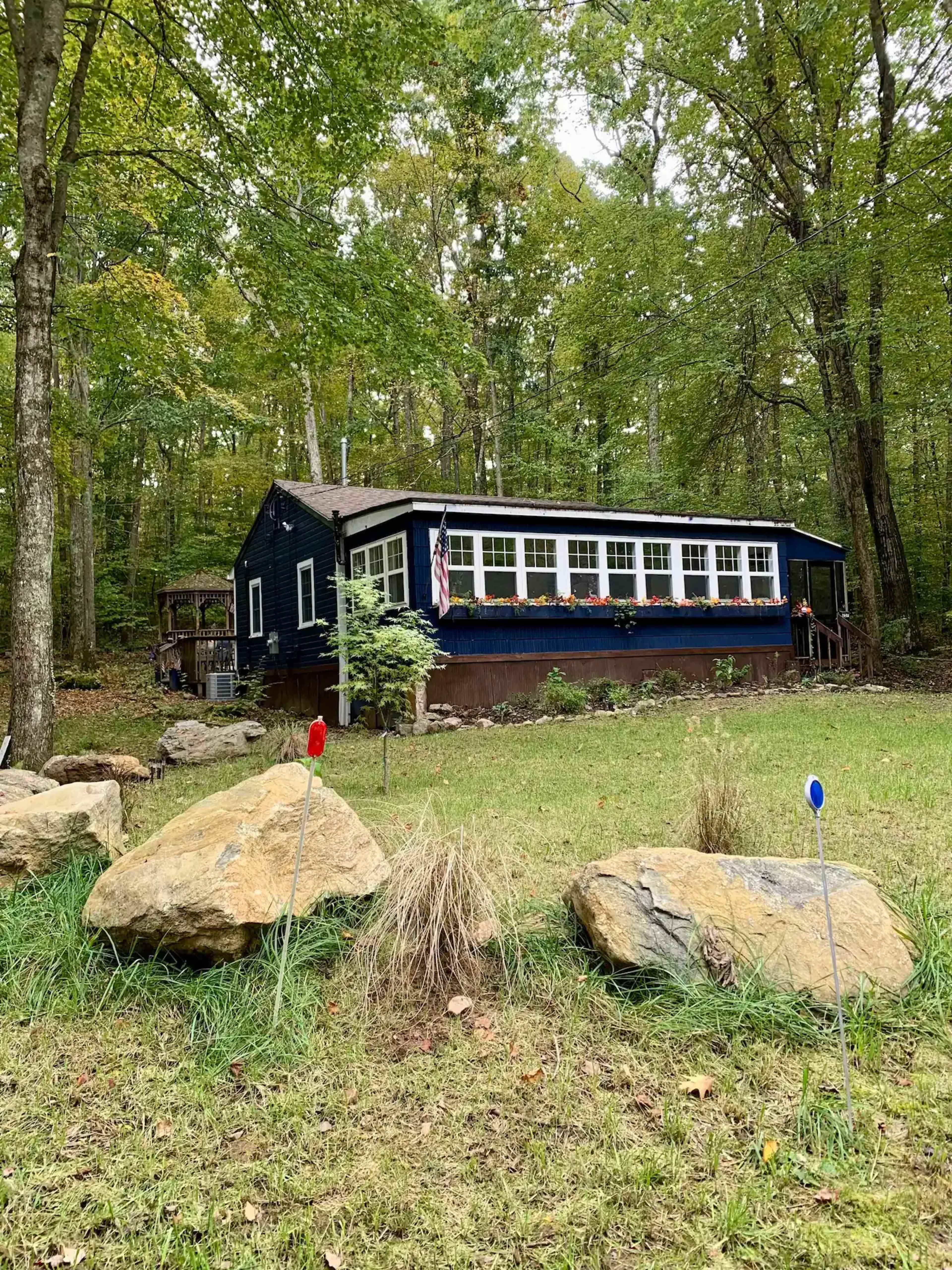 A blue house with a porch and large windows is nestled among trees in a grassy area, surrounded by boulders and decorative markers.