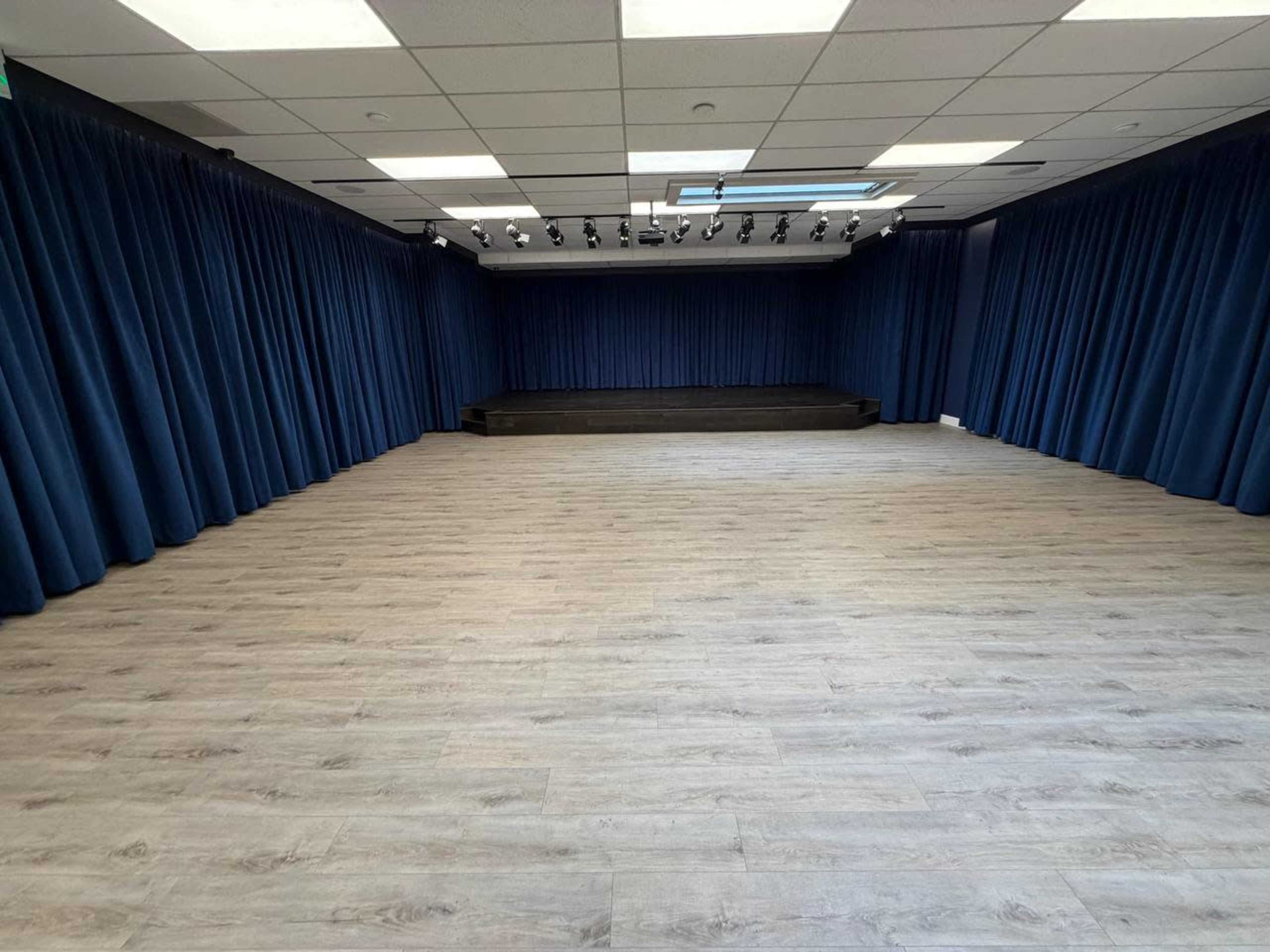The image shows a spacious room with wooden flooring and blue curtains lining the walls, featuring a raised platform at one end.