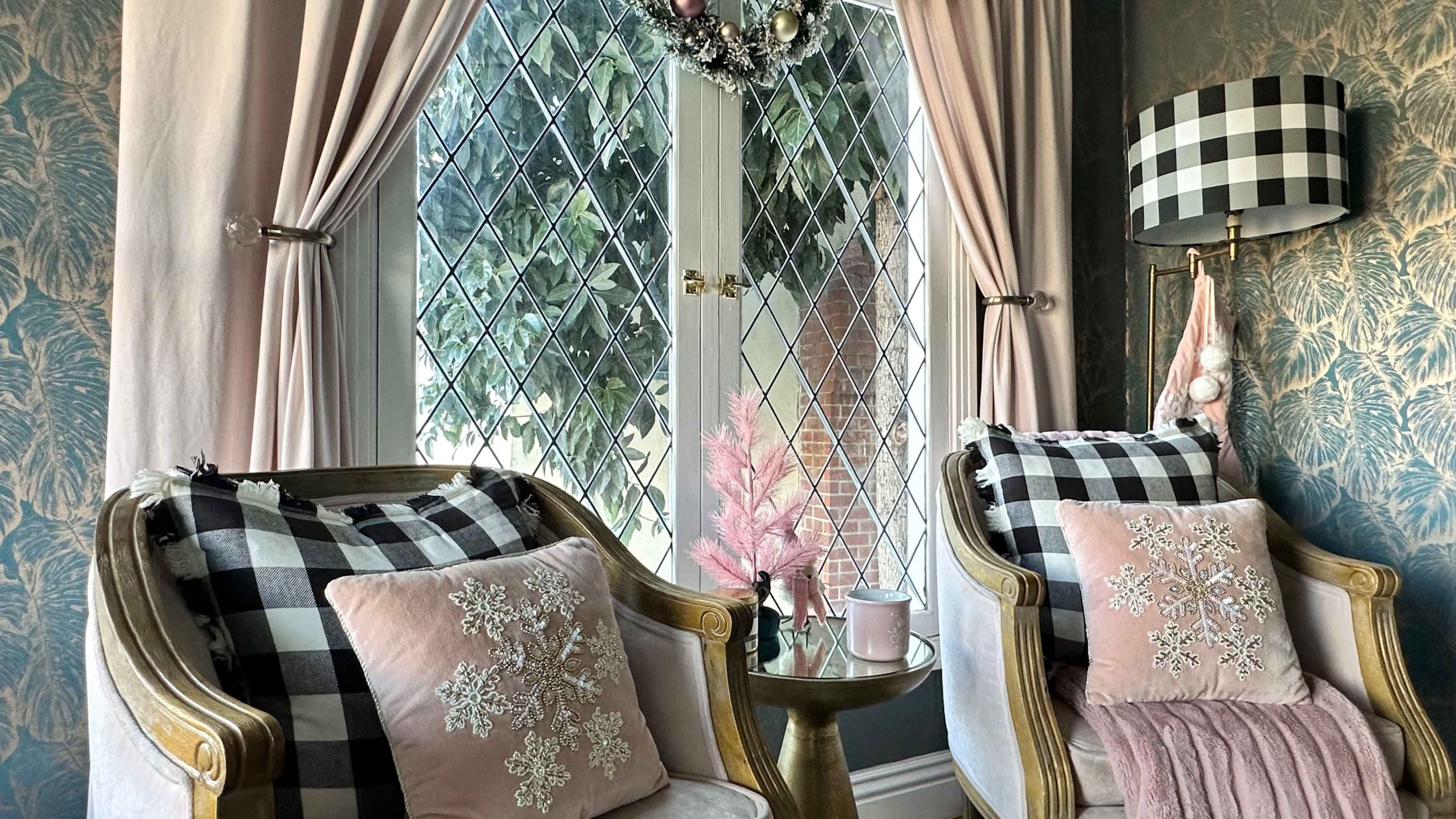 The image shows a cozy seating area with two upholstered chairs, a decorative table, and a window adorned with curtains, a wreath, and a lamp.