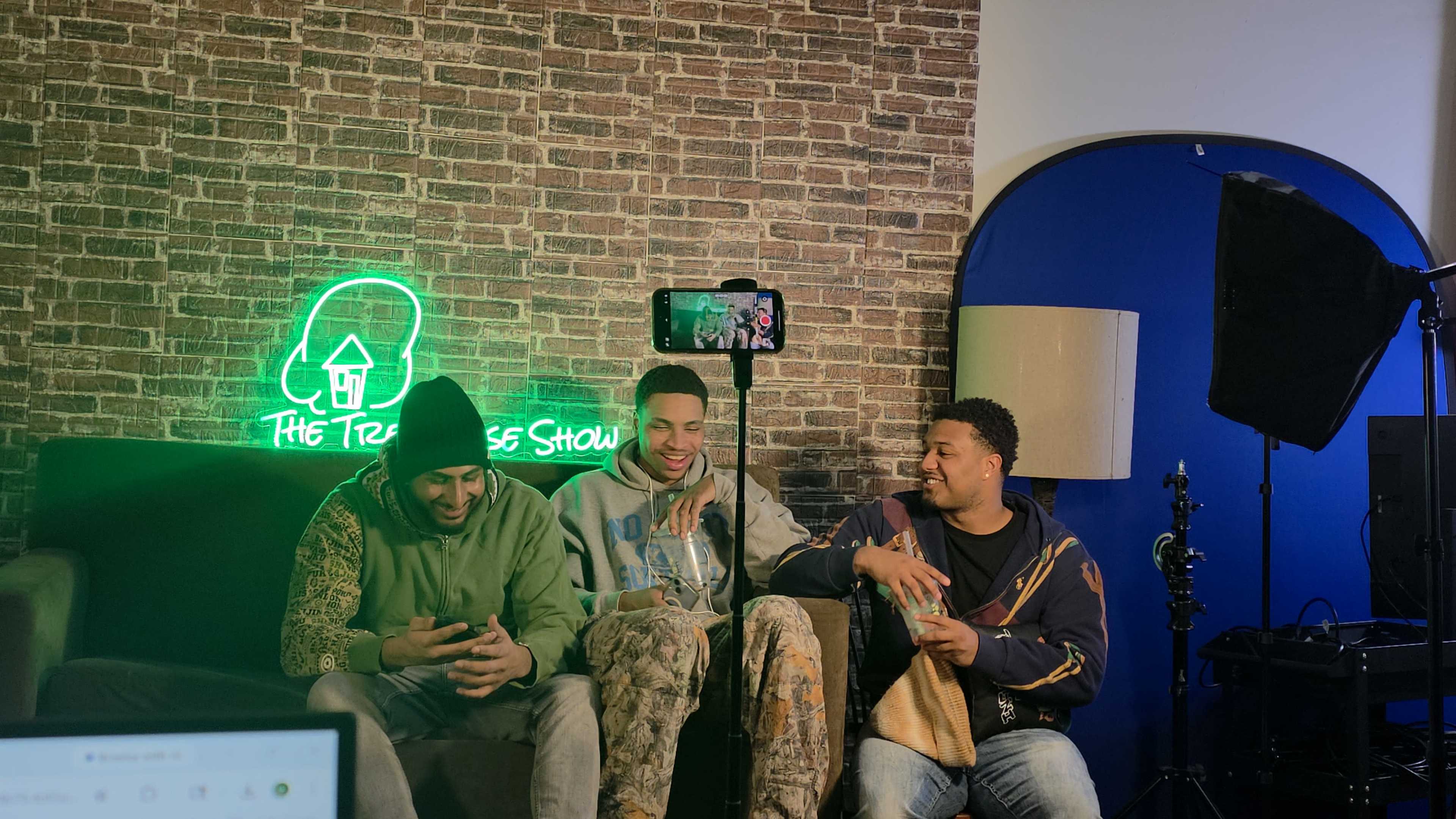 Three young men are sitting on a couch, engaged in conversation, with a smartphone on a tripod capturing the moment and a neon sign in the background that reads "The Treehouse Show."
