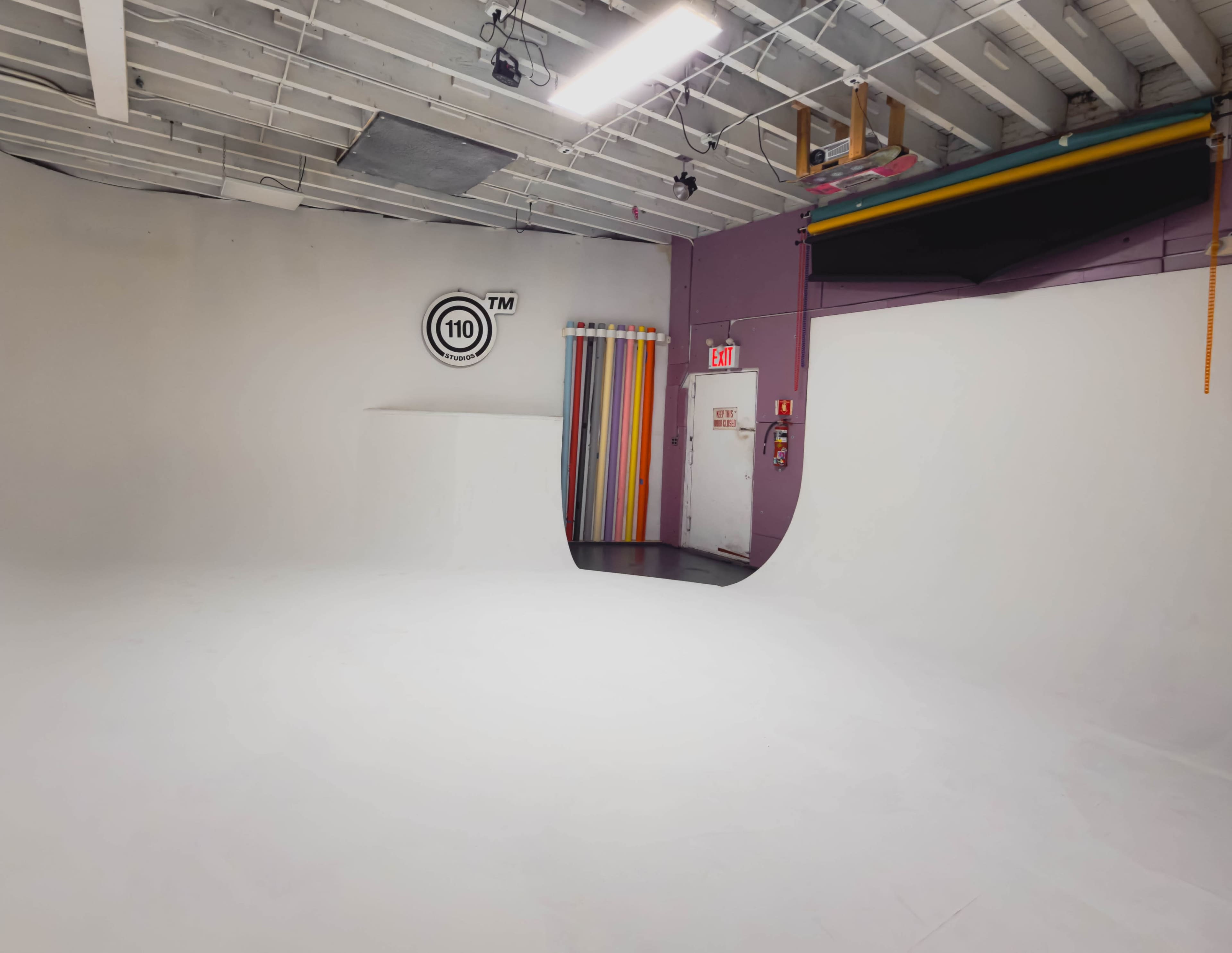 The image shows a spacious, white studio with a curved backdrop, colorful curtains, and a door leading to another area.