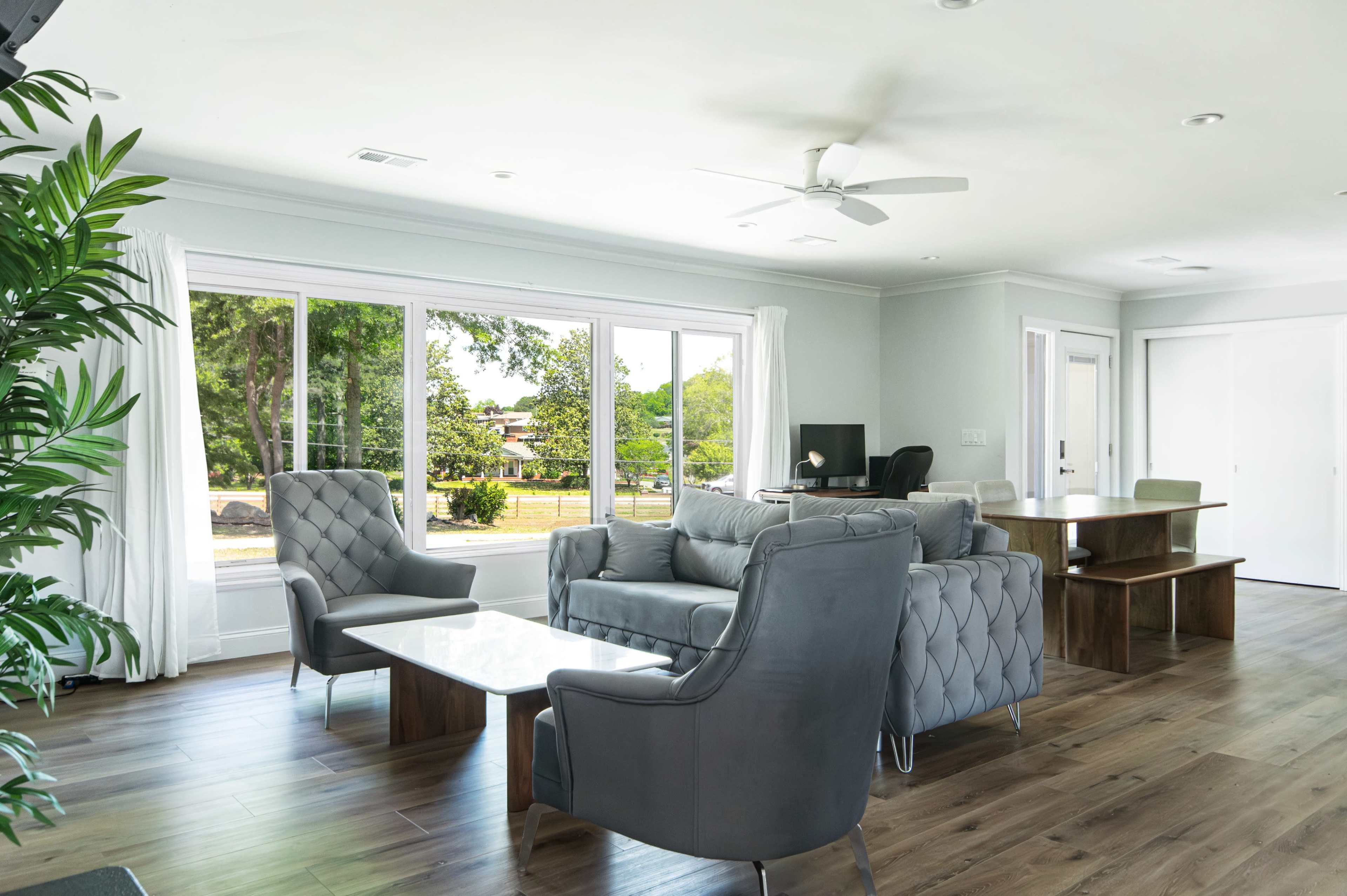 The living area features a modern seating arrangement with gray chairs and a sofa, large windows allowing natural light, and a wooden floor.