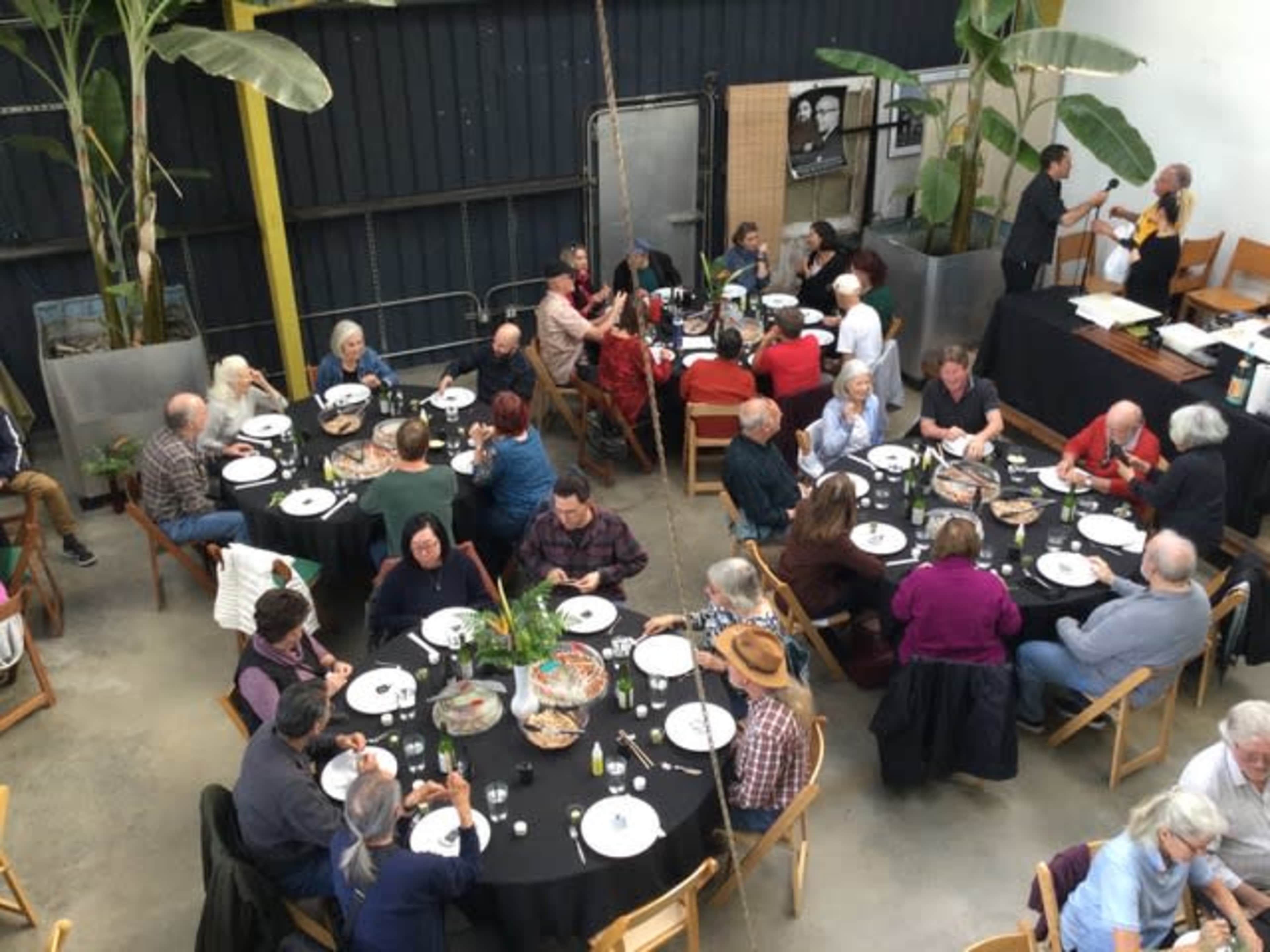 A large group of people sits at round tables in a spacious dining area, enjoying a meal while surrounded by potted plants.