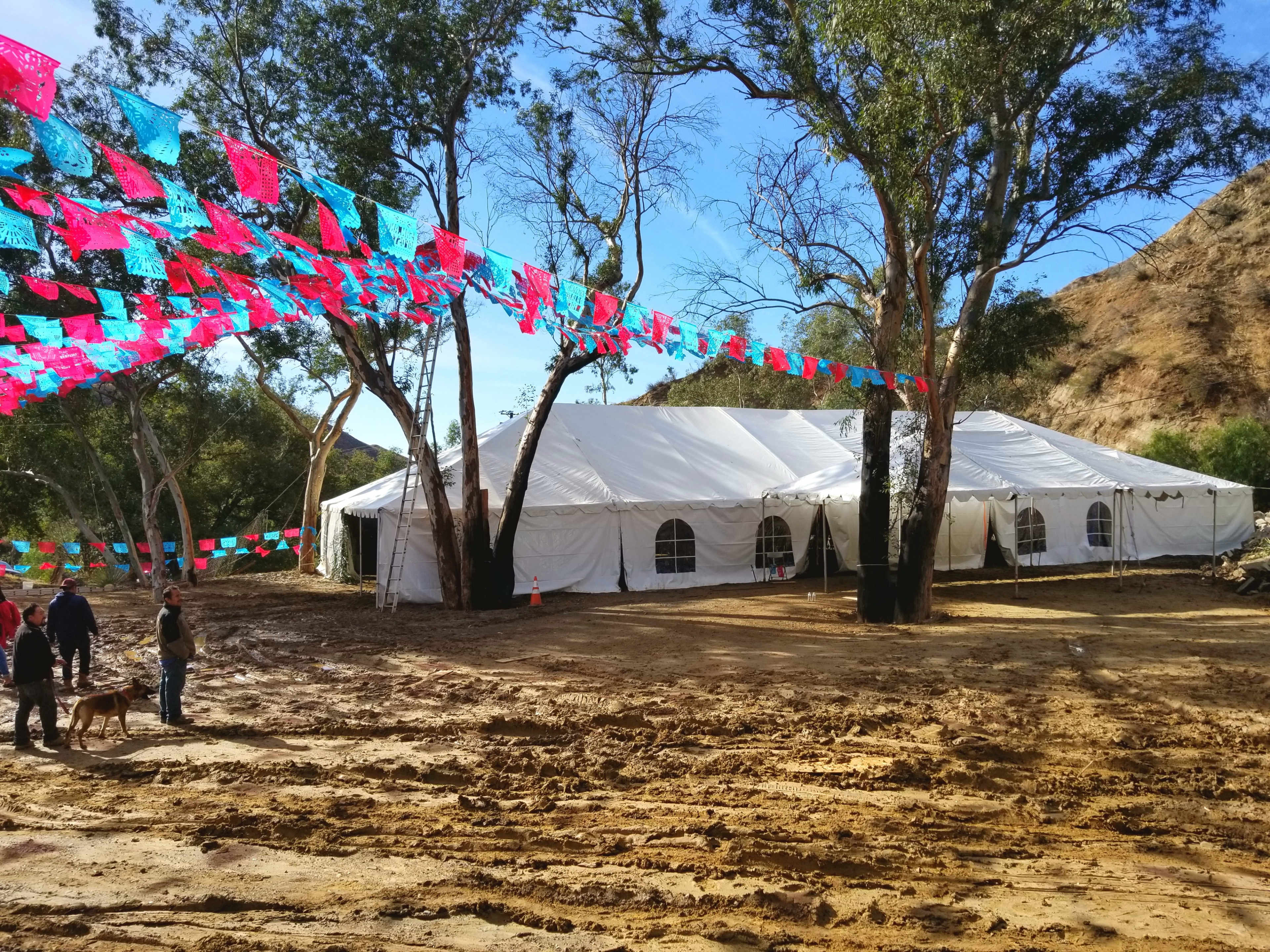 A large white tent is set up in a muddy clearing, surrounded by trees and colorful decorative banners overhead.