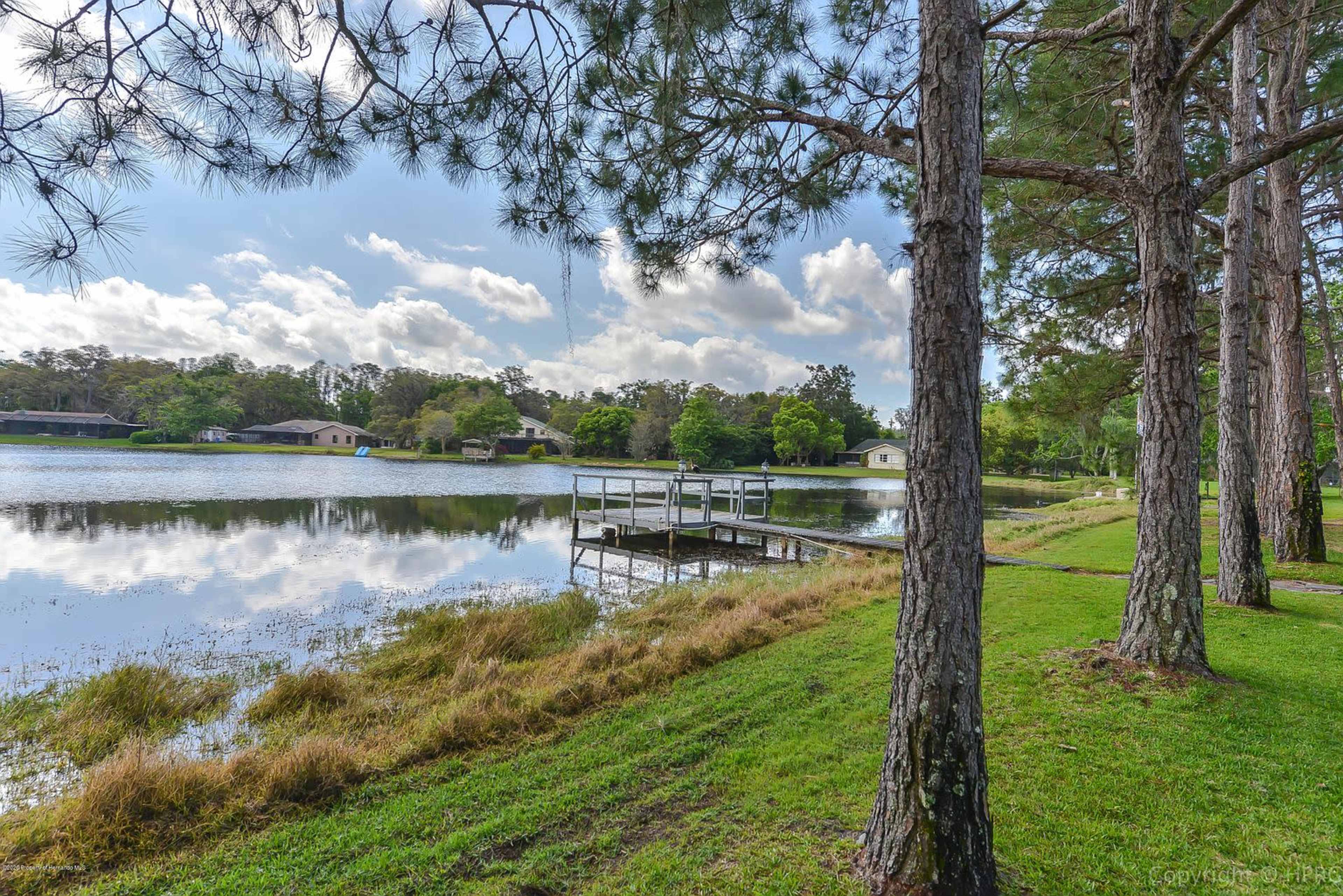 A calm lake bordered by trees and a small dock, with houses visible in the background.