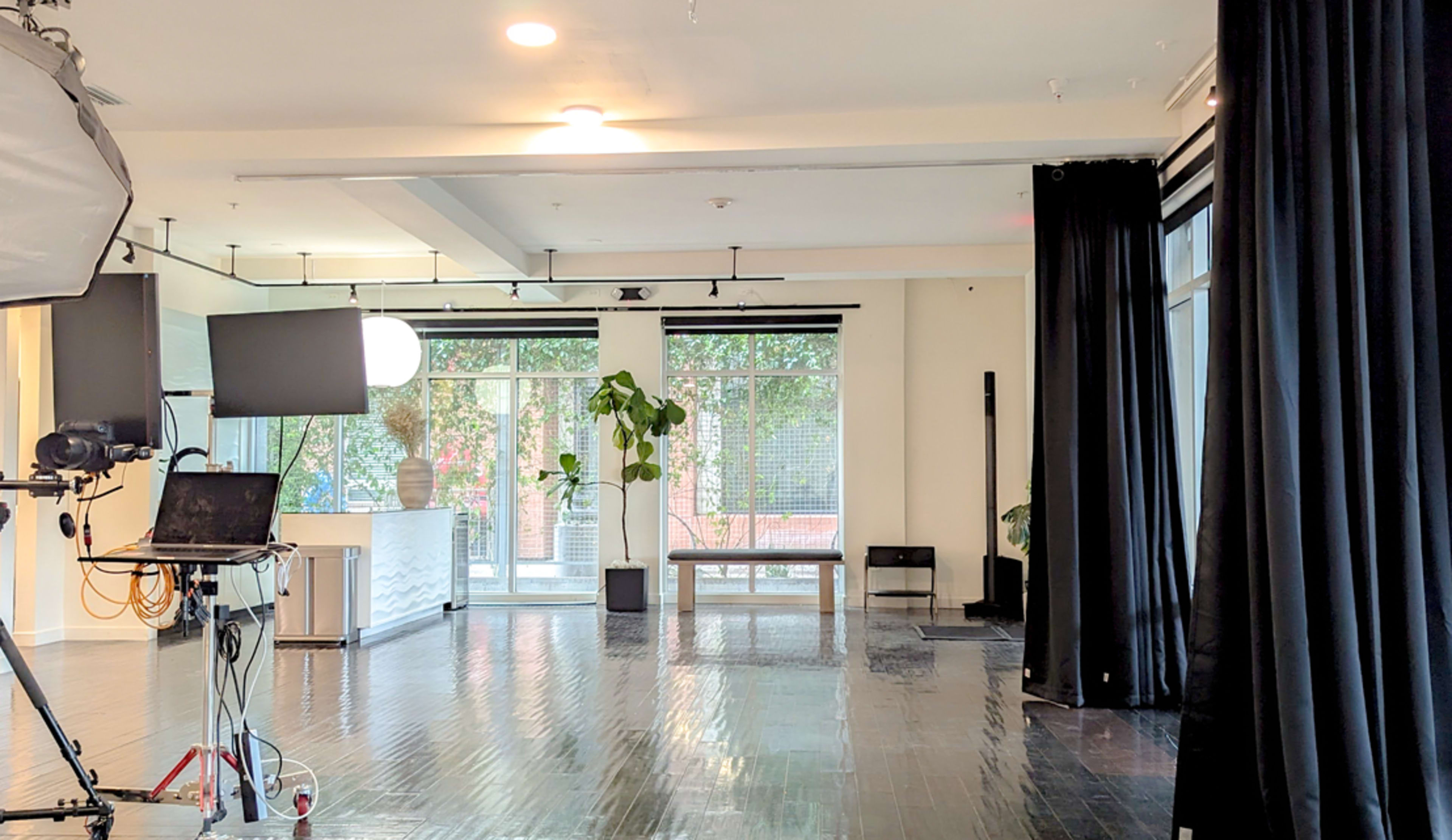 A bright, open studio space with large windows, dark curtains, and a video setup including a camera and monitor.