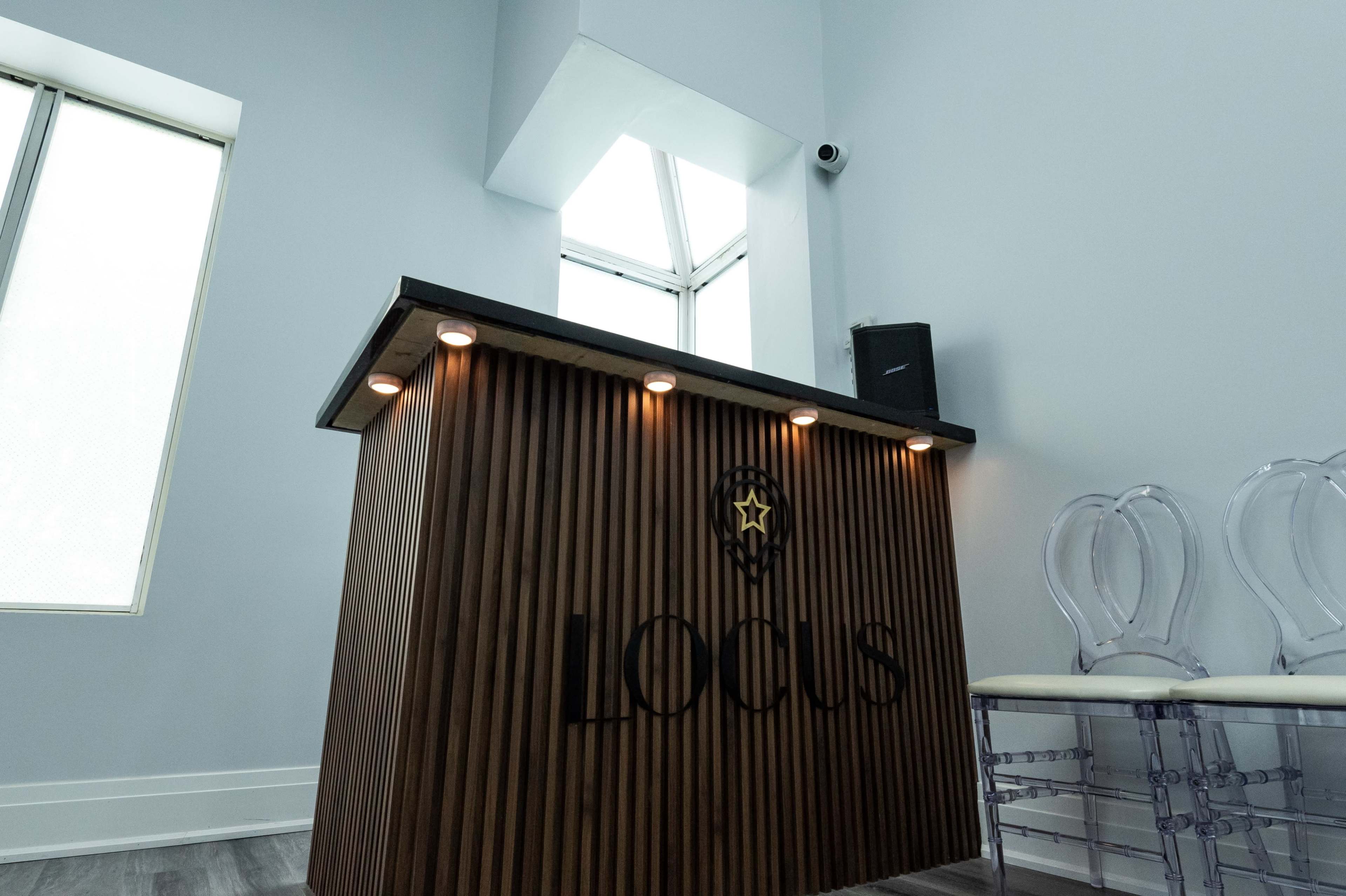 A modern reception desk with a wood panel design and the word "Locus" displayed, positioned next to clear acrylic chairs and a window above.