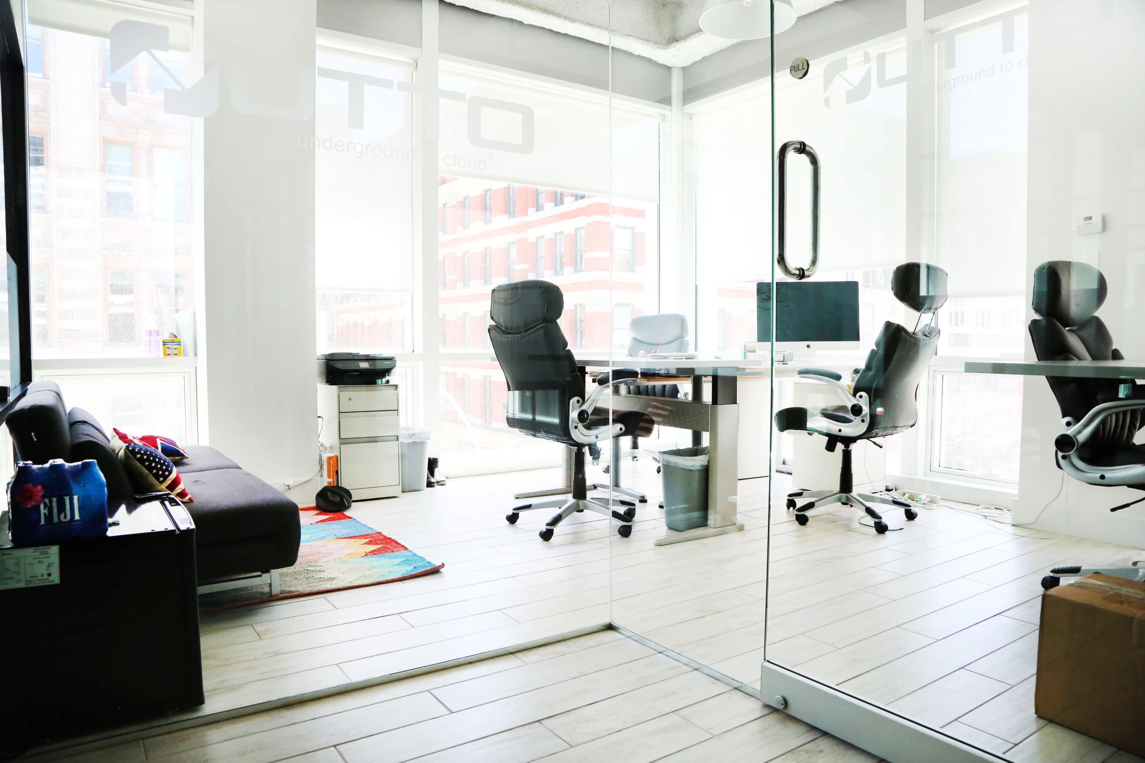 A modern, glass-walled office space with a couch, a desk with a computer, and several ergonomic chairs.