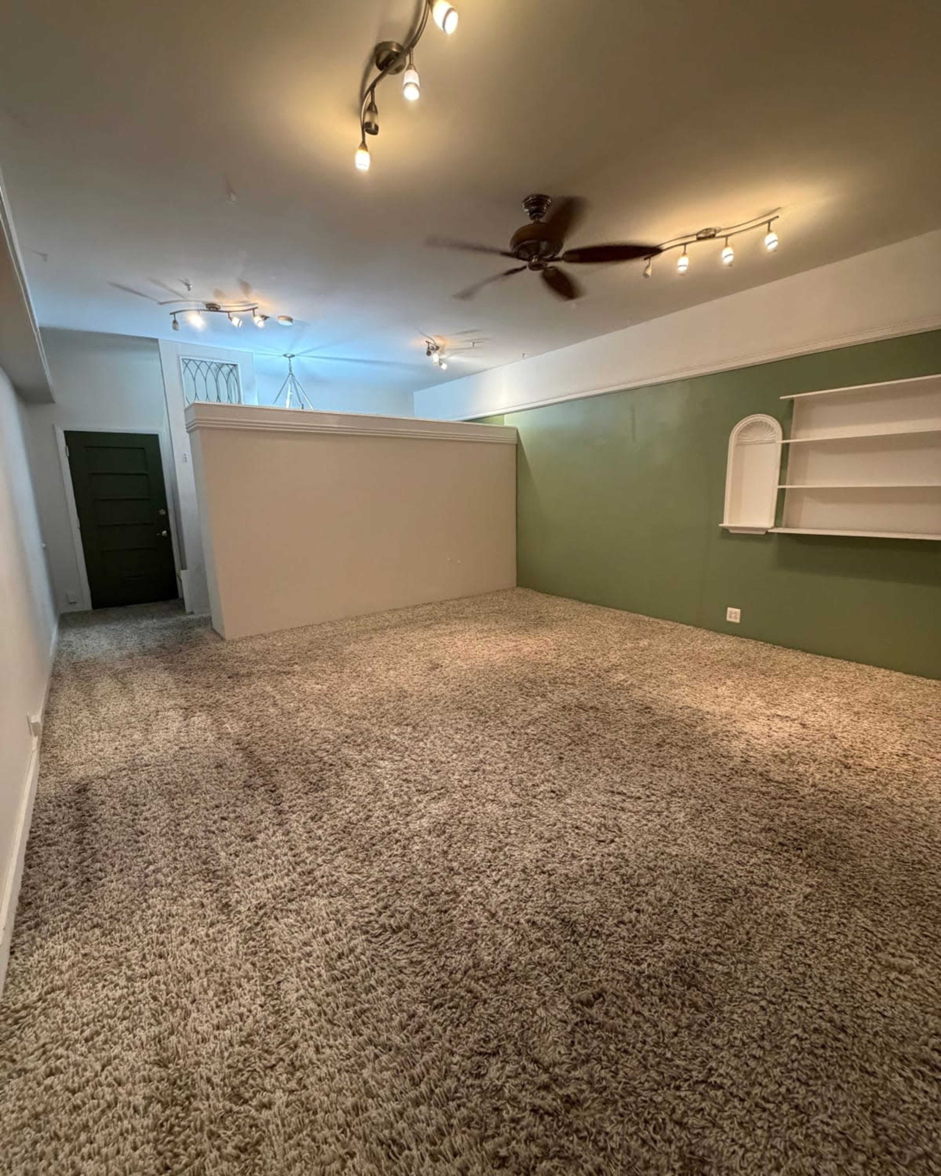 The image shows a spacious, carpeted room with green walls, a ceiling fan, and wall-mounted shelves, along with an entrance visible on the left side.