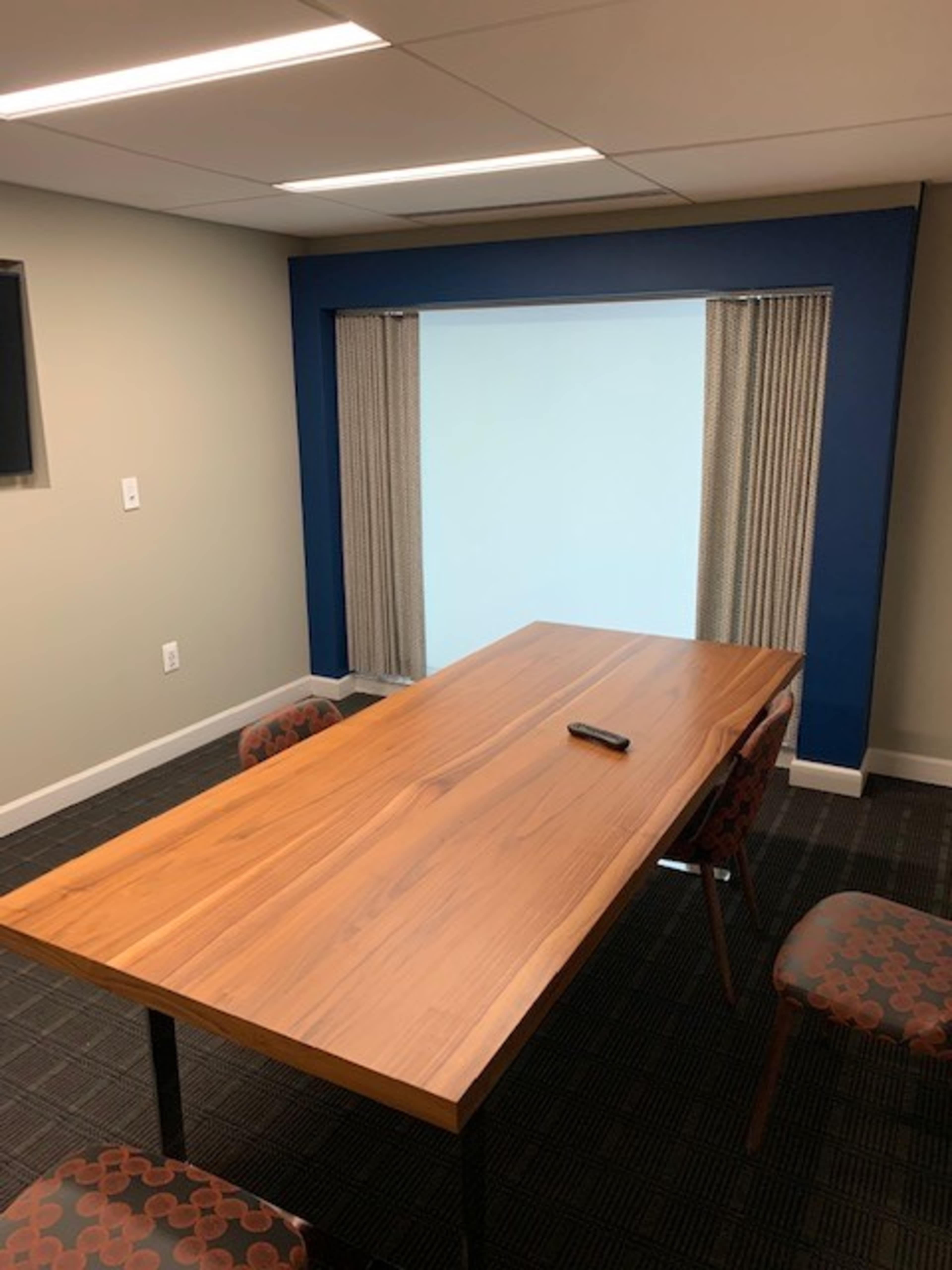 A wooden conference table is positioned in a small meeting room with a window covered by curtains.