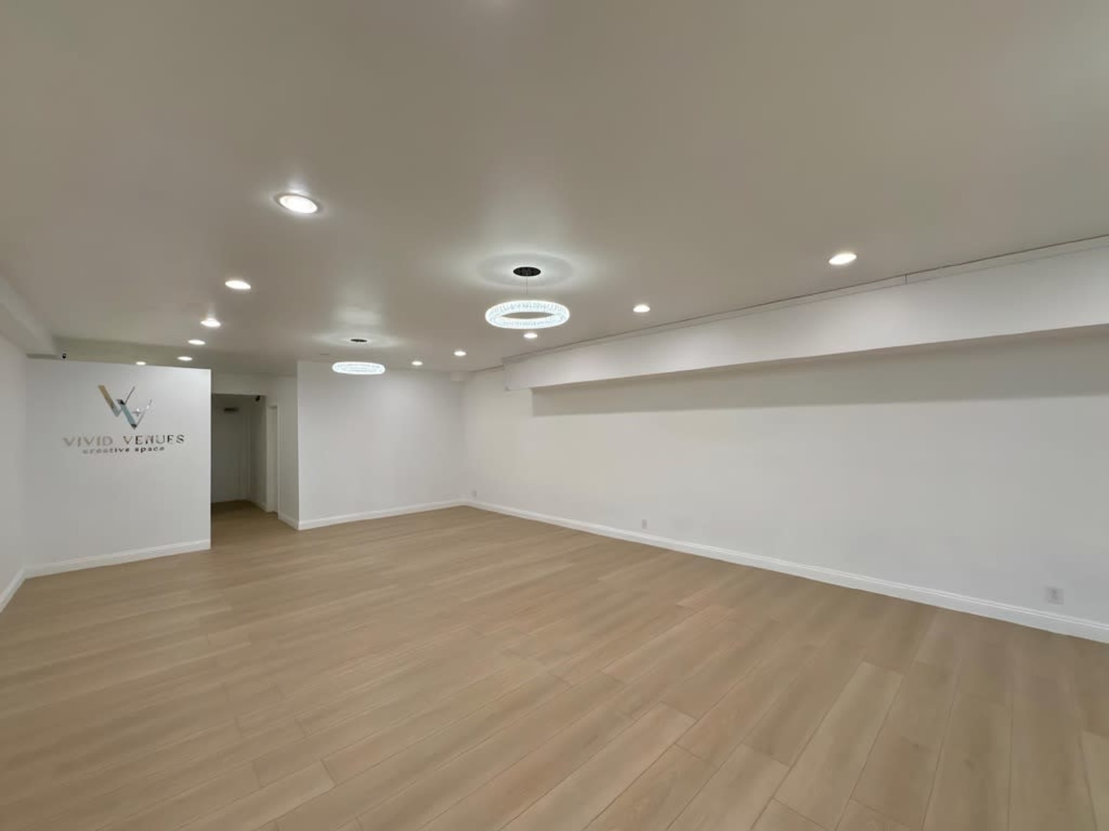 The image shows a spacious, empty room with wooden flooring and white walls, featuring circular ceiling lights and a logo on one wall.