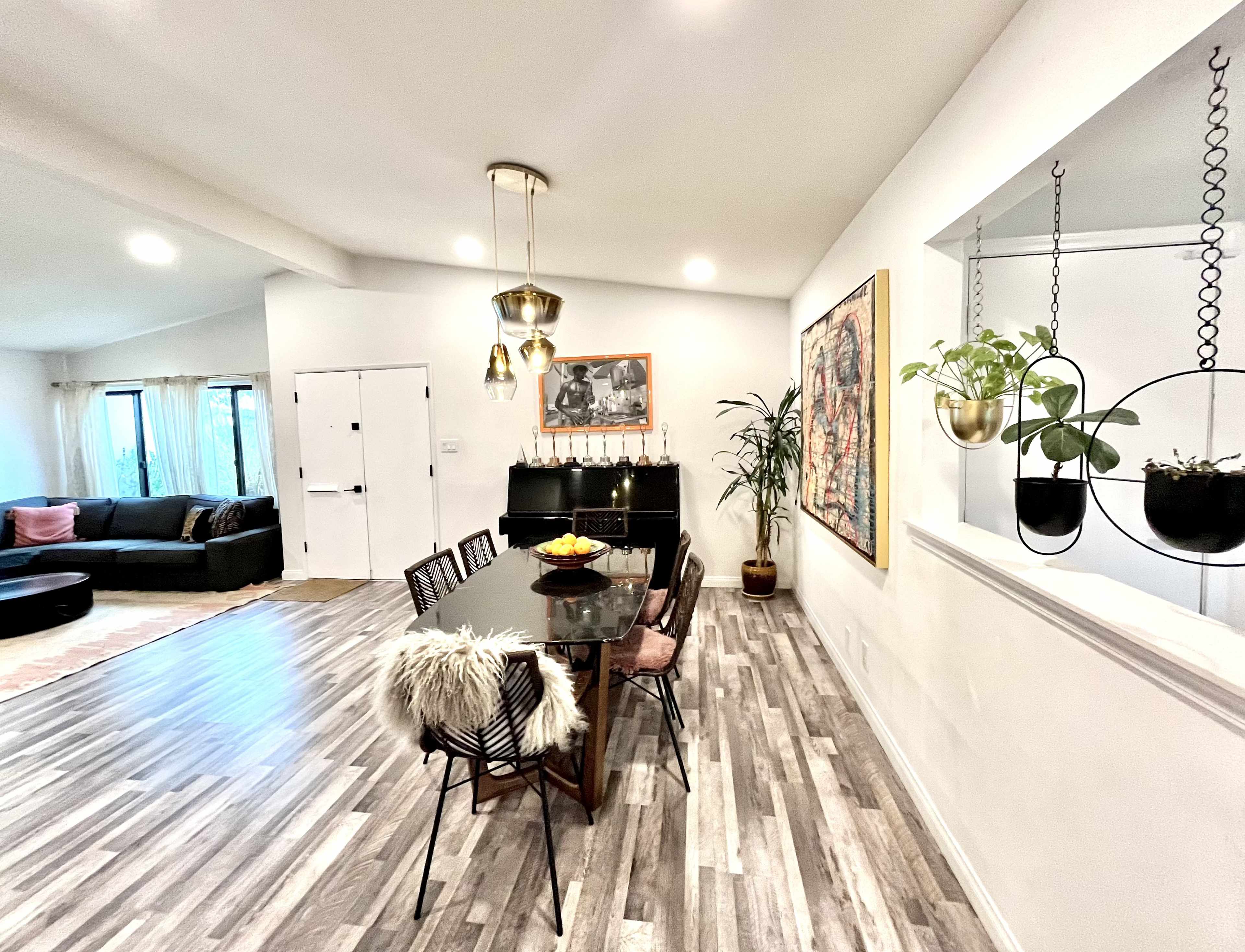 A modern dining area with a black table surrounded by chairs, a potted plant, and artwork on the wall, adjacent to a living space featuring a large sofa.
