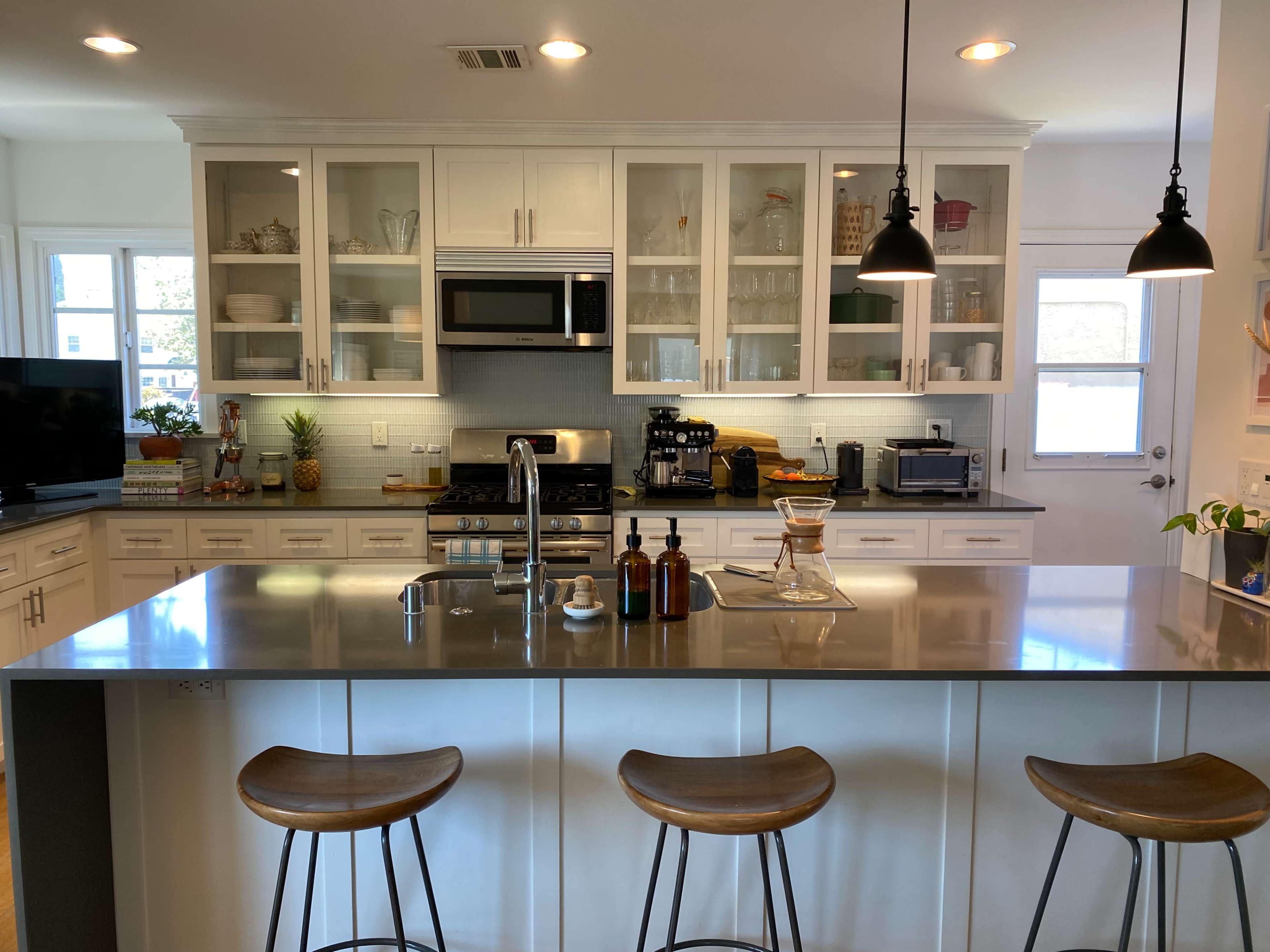 A modern kitchen with a central island, three bar stools, and glass-front cabinets displaying various kitchenware.