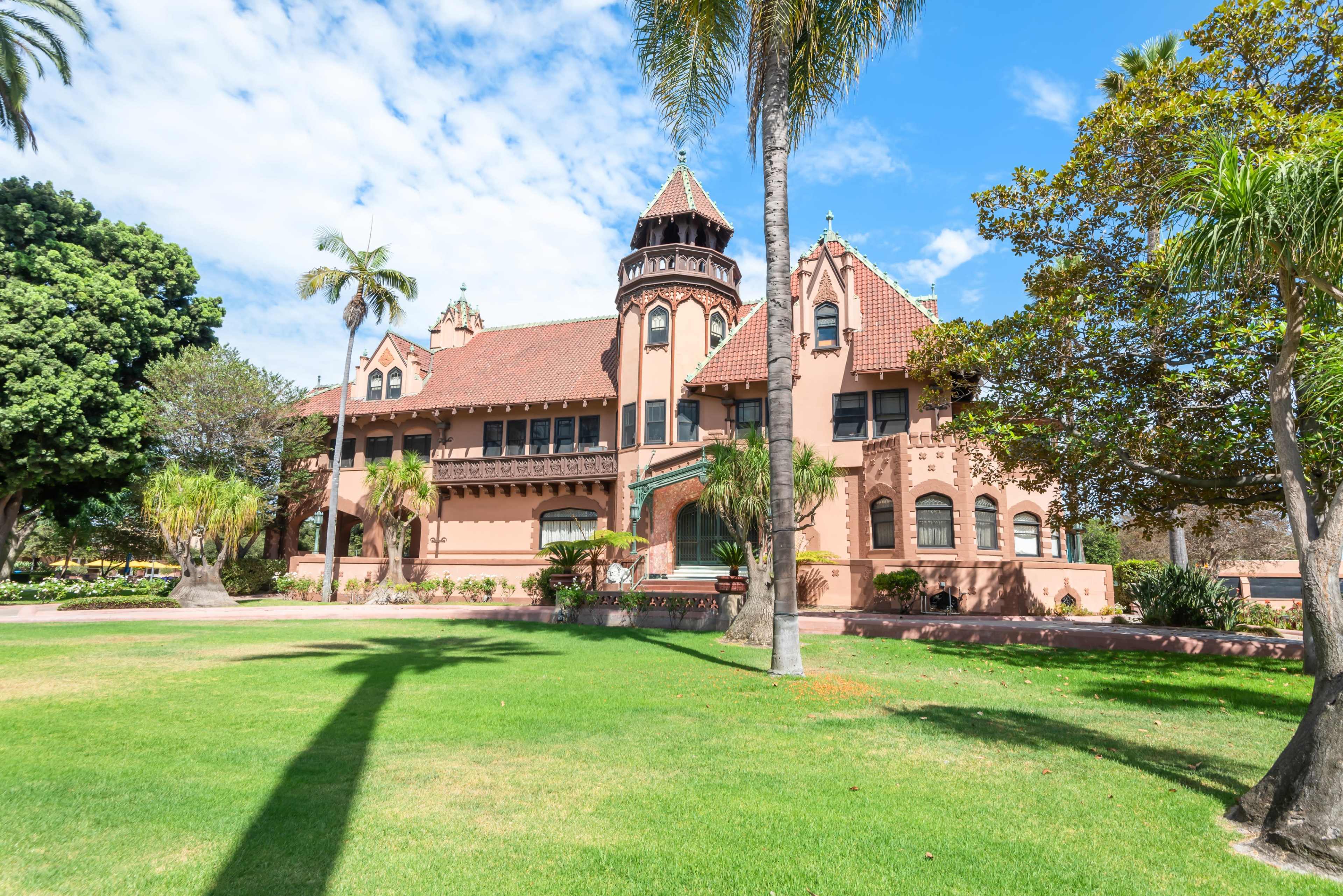 The image shows a large, historic mansion with a distinctive tower, surrounded by palm trees and lush green grass.
