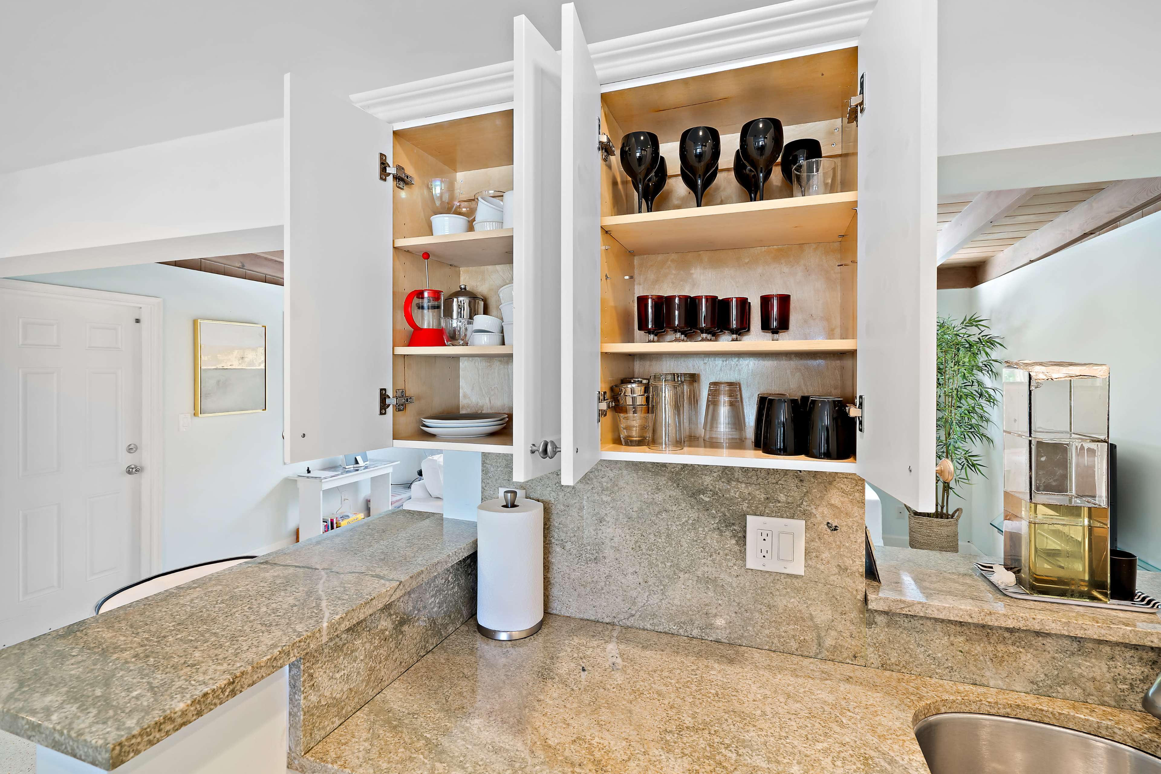 The image shows a kitchen with open cabinets displaying various glassware and dishes, along with a granite countertop and a doorway in the background.