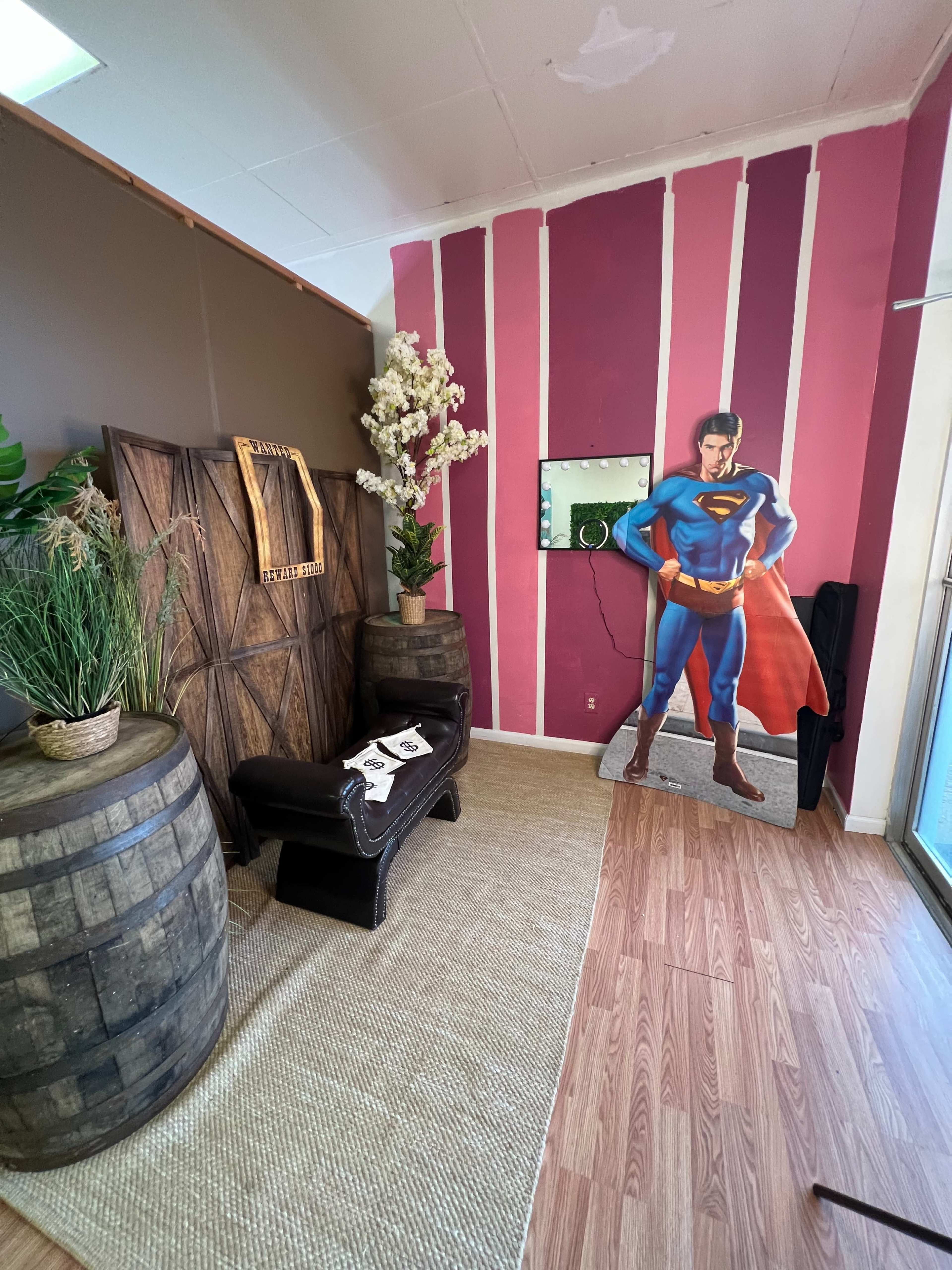 A room with a Superman cutout, a bench, and decorative elements like plants and barrels.