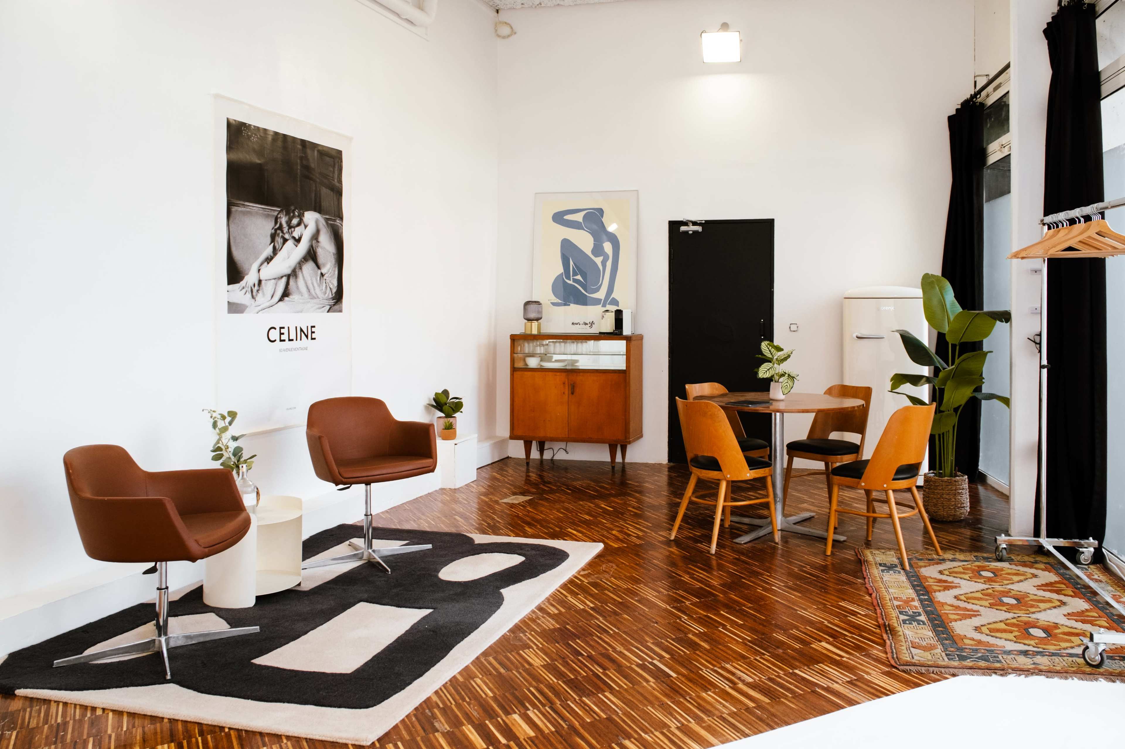 The image shows a modern interior with two brown chairs, a wooden dining table, a vintage cabinet, and potted plants against white walls and a hardwood floor.