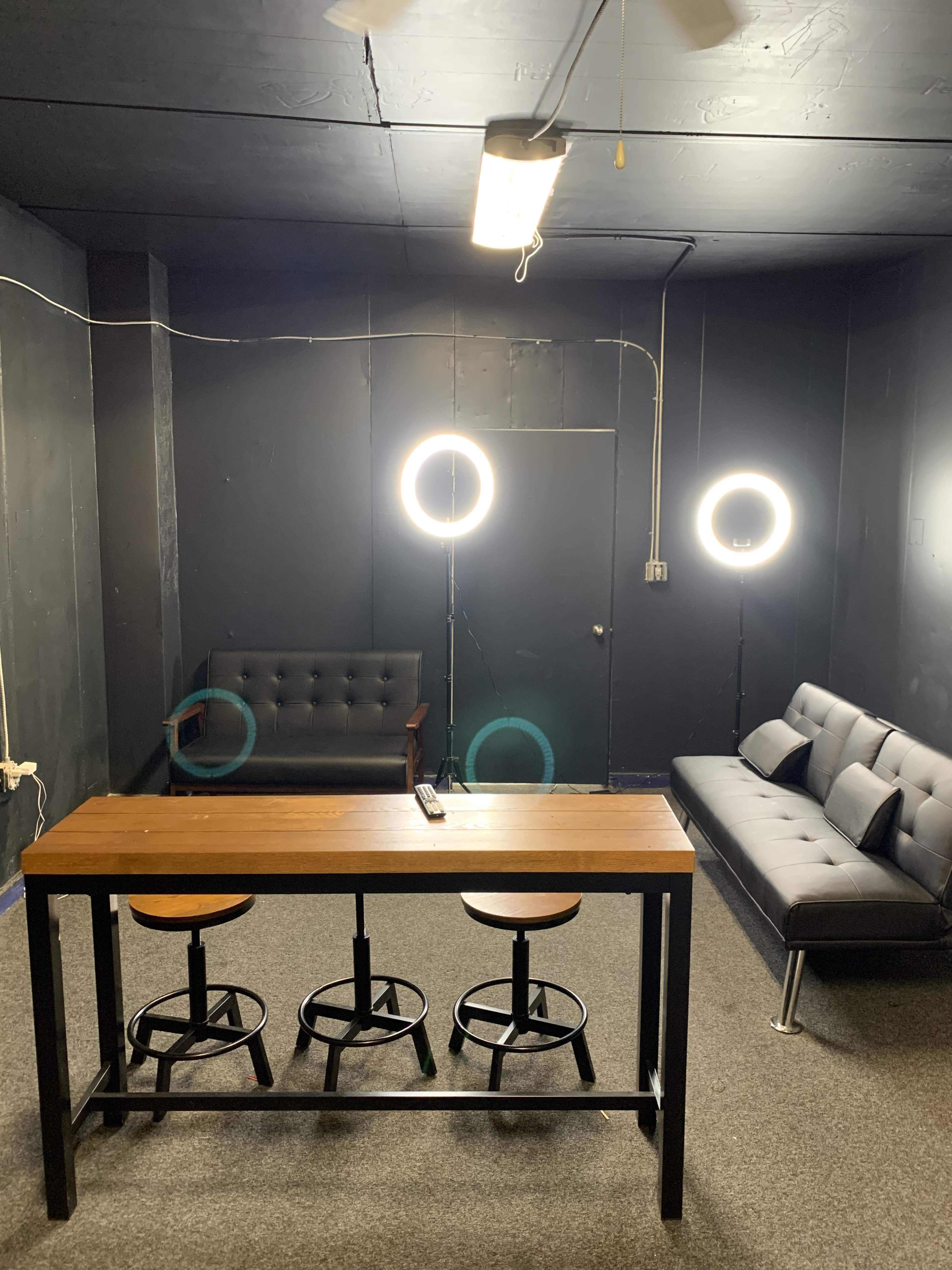 A dimly lit room features a table with three stools, a black couch, and two ring lights illuminating the space.