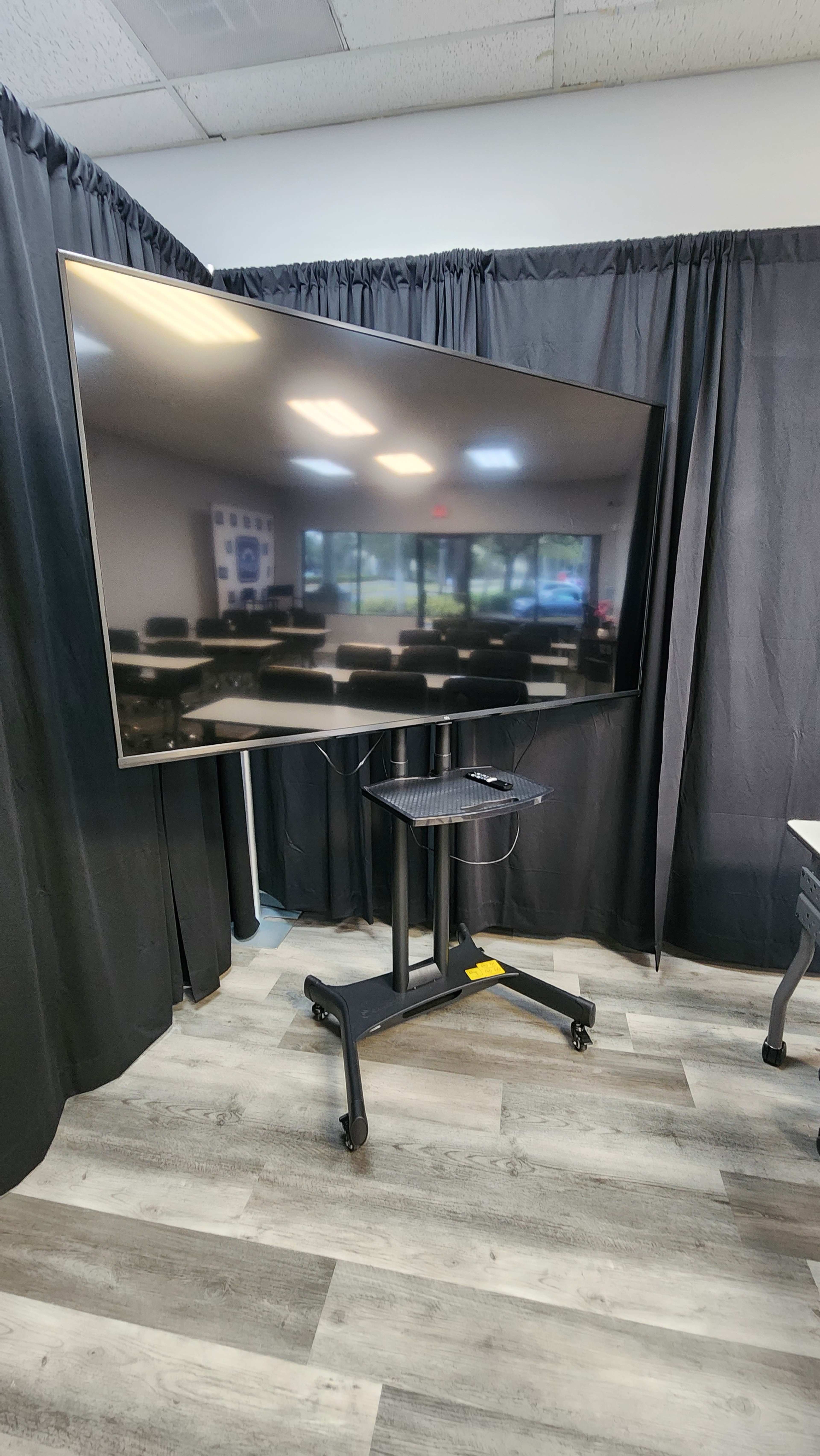A large flat-screen monitor is mounted on a mobile stand in a room with black curtains and a view of chairs and tables in the background.