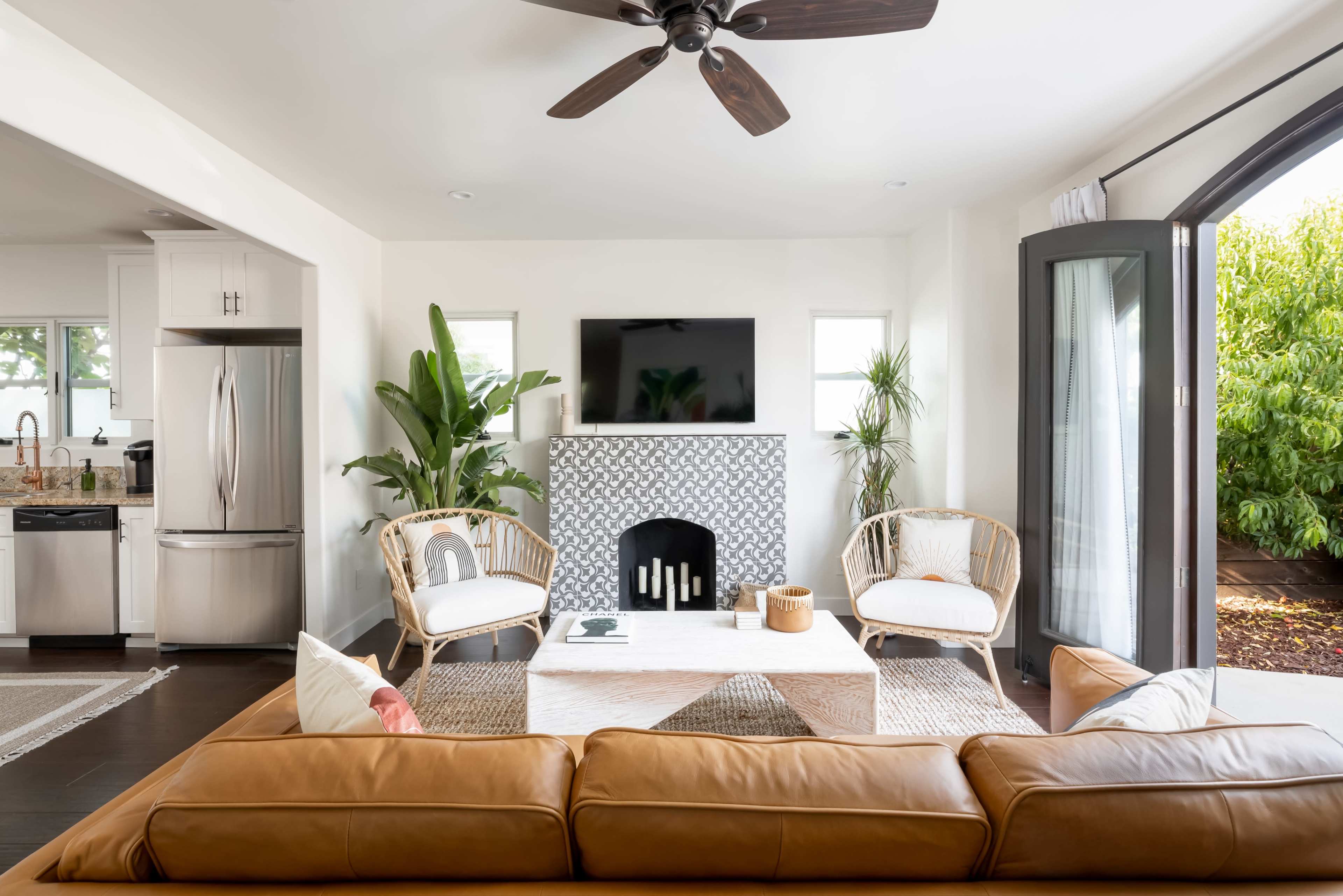 A modern living room features a brown leather couch, two rattan chairs, a coffee table, and a patterned fireplace, with a television mounted on the wall and large windows opening to greenery outside.