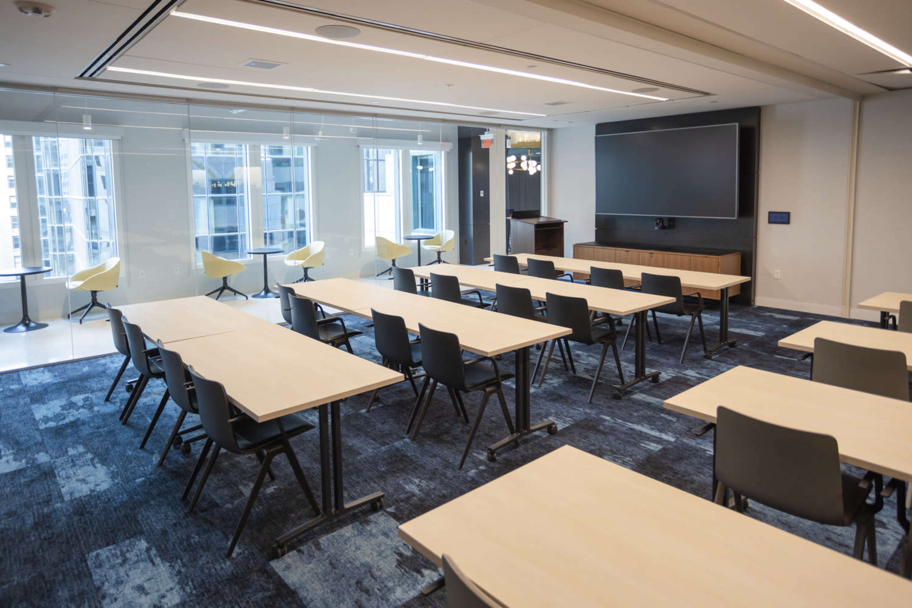 A modern classroom setup with rows of tables and chairs, large windows providing natural light, and a presentation screen at the front.