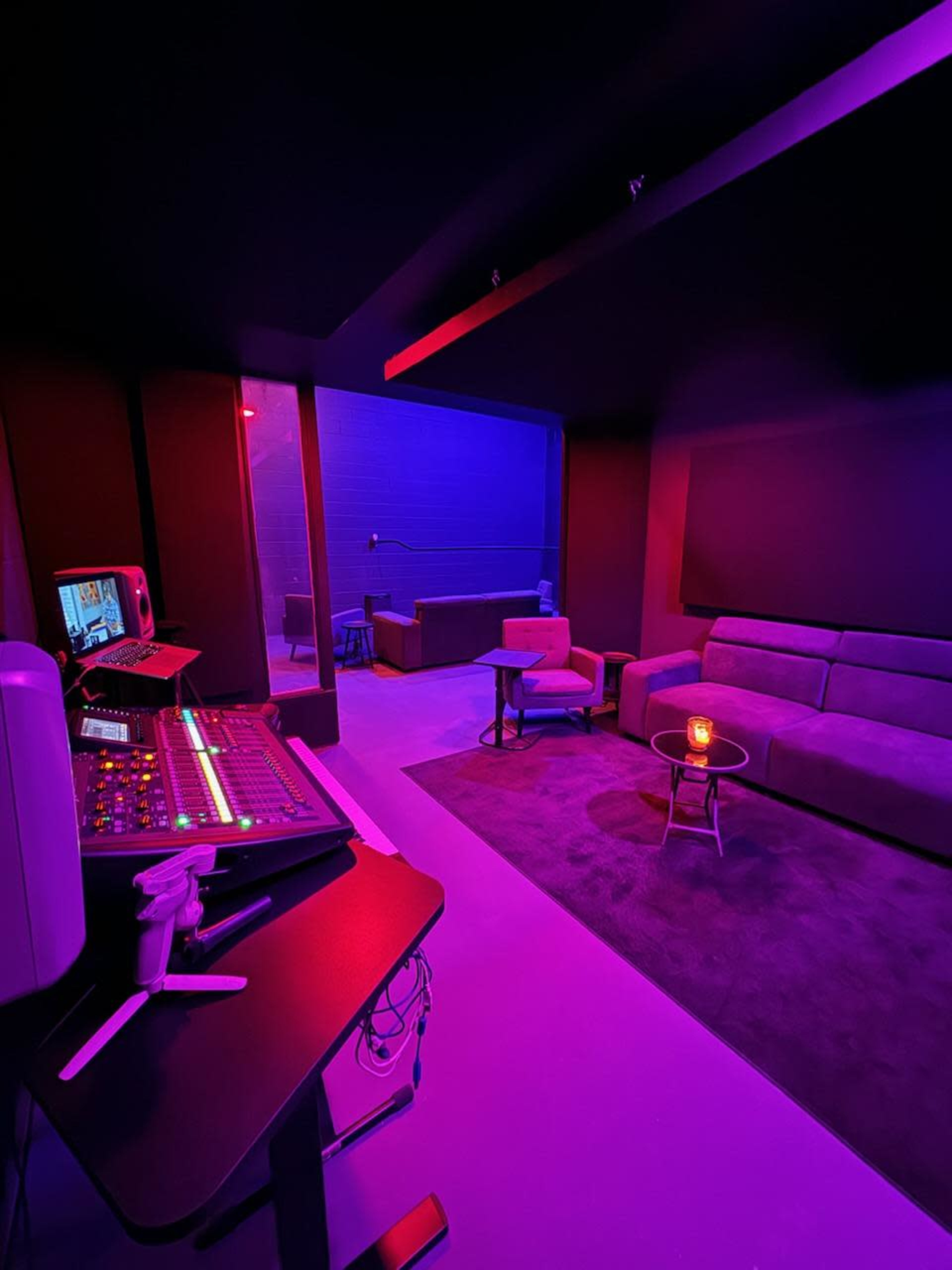 The image shows a dimly lit recording studio with a control panel on the left and a seating area featuring a couch and a small table in the background.