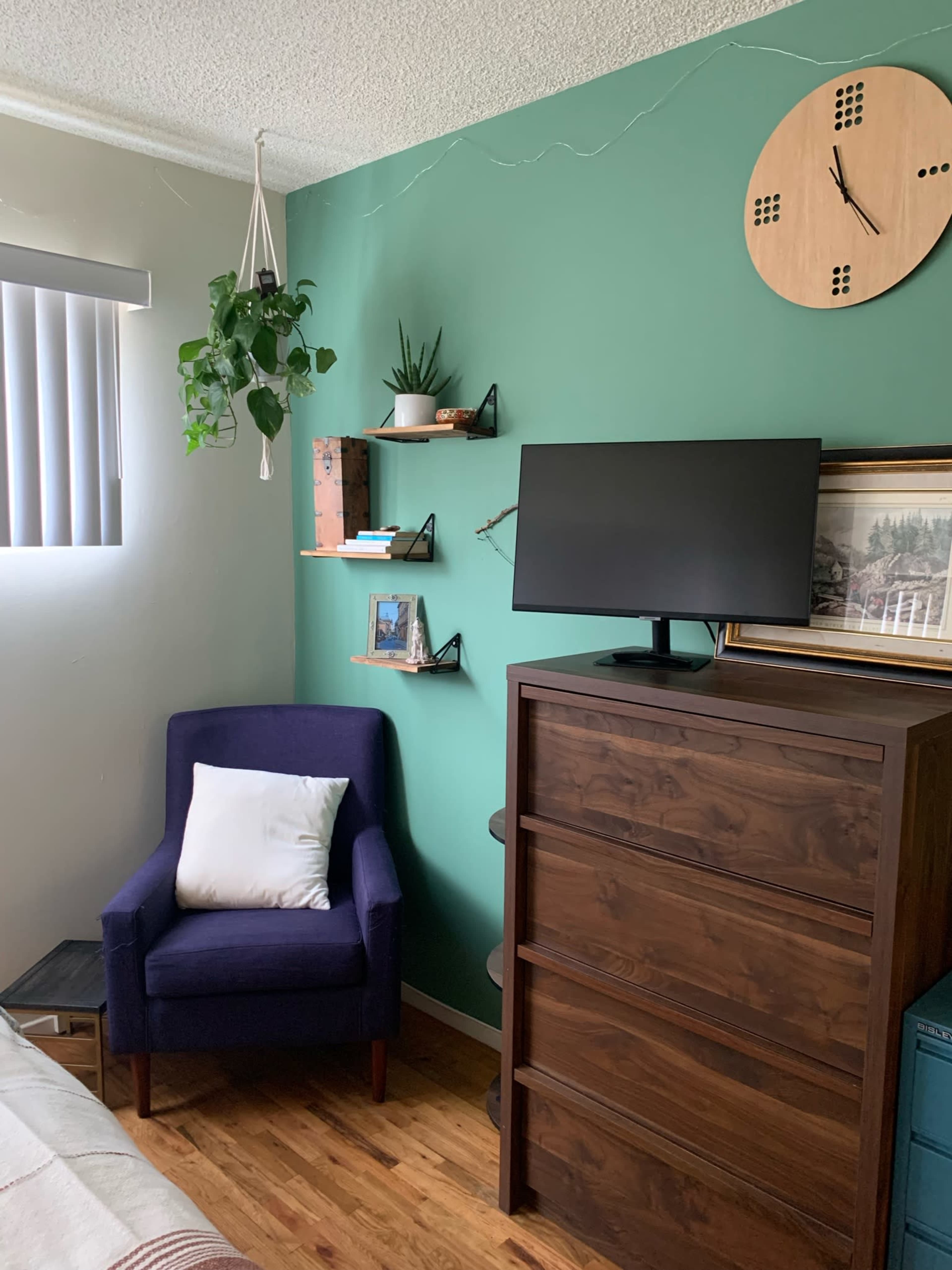A cozy room features a dark wood dresser, a purple armchair with a white pillow, and a wall adorned with plants and shelves, alongside a wall clock and a television.