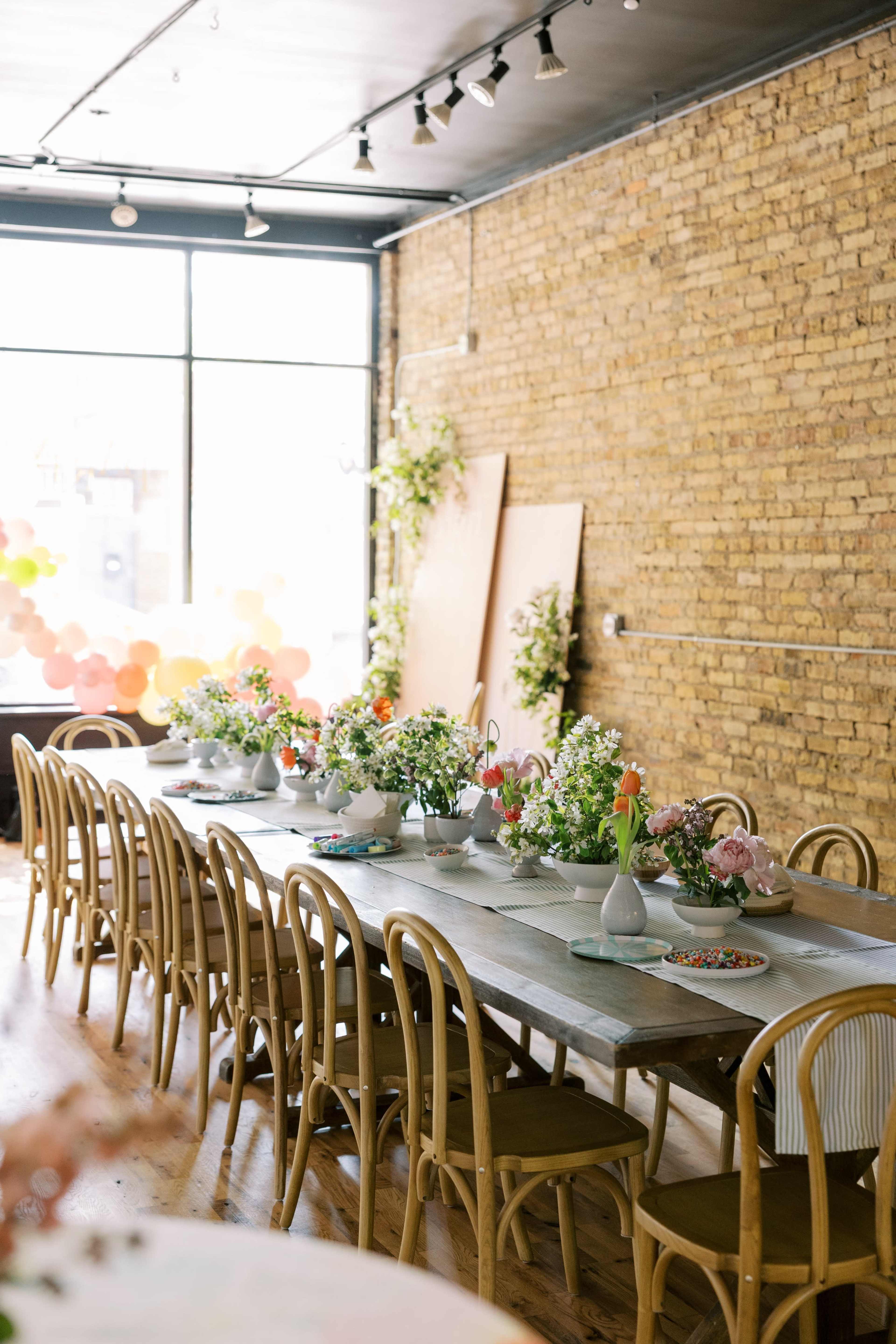 A long wooden table set with floral arrangements and tableware, situated in a brightly lit room with exposed brick walls.