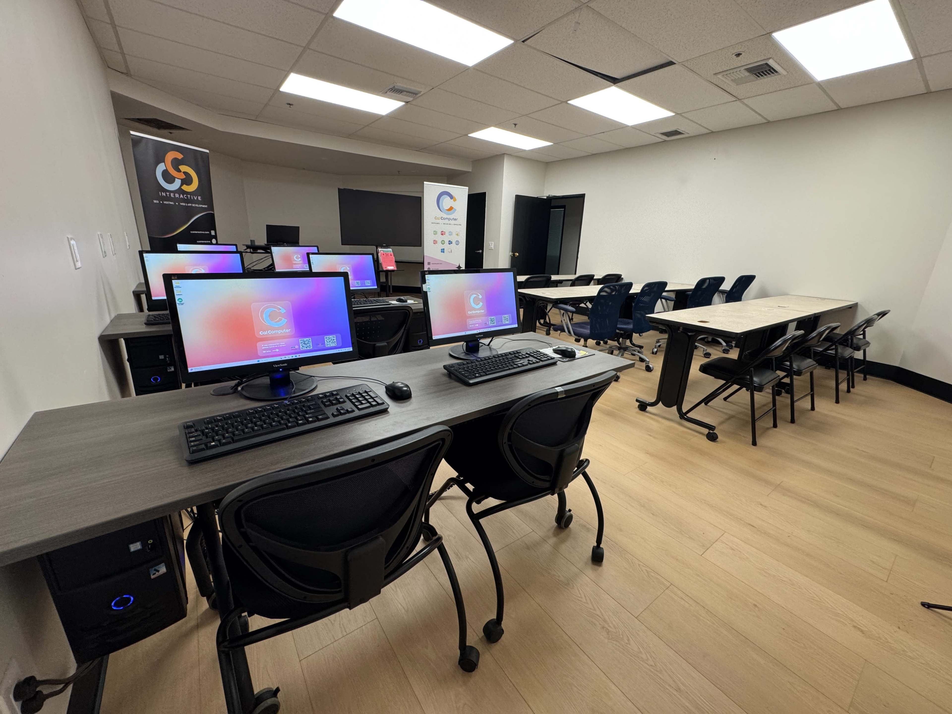 The image shows a modern classroom equipped with desktop computers and desks, featuring a series of blue rolling chairs and a bulletin board.