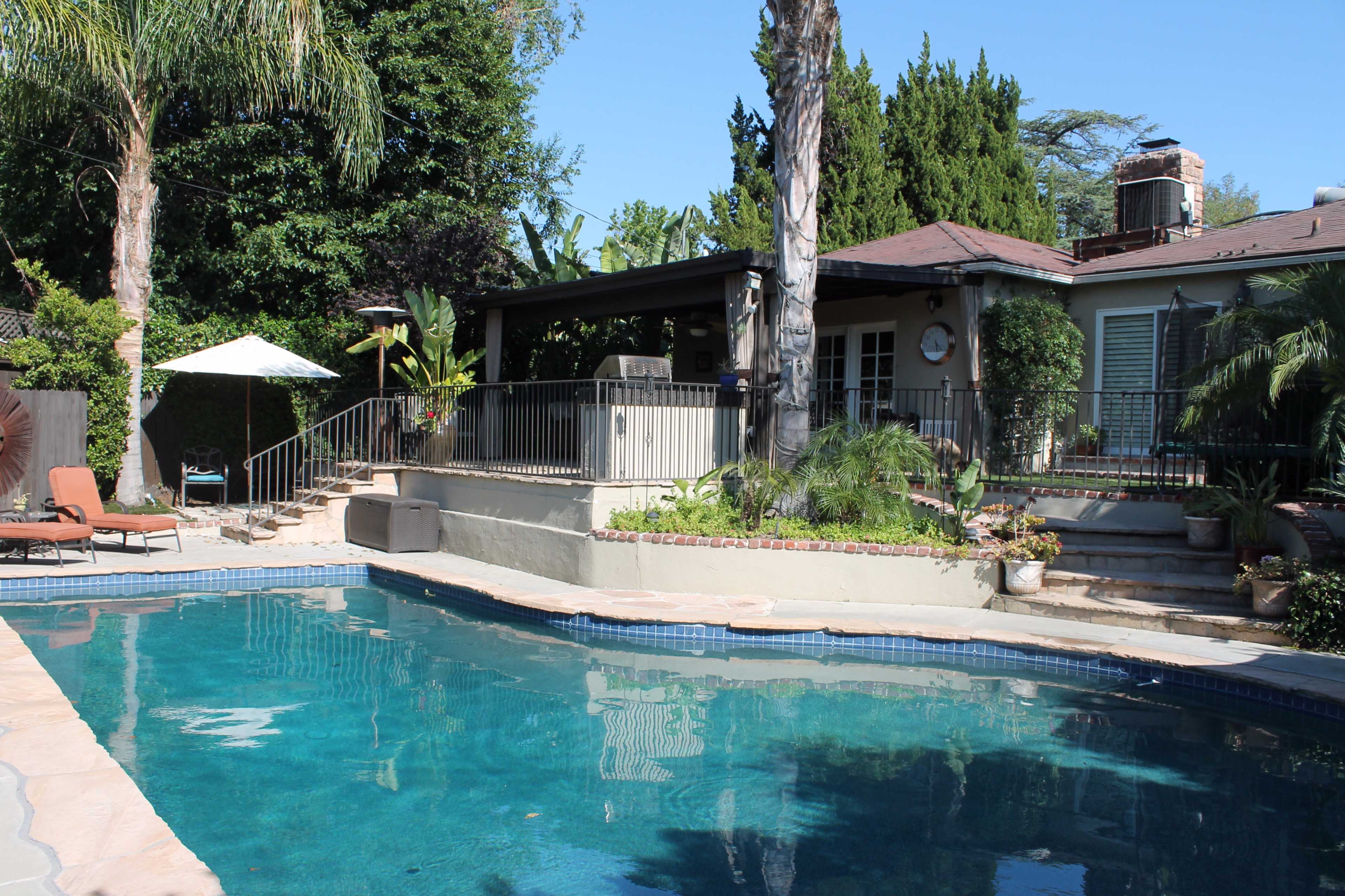 The image shows a backyard with a swimming pool, surrounded by tropical plants and a patio area with lounge chairs and an umbrella.