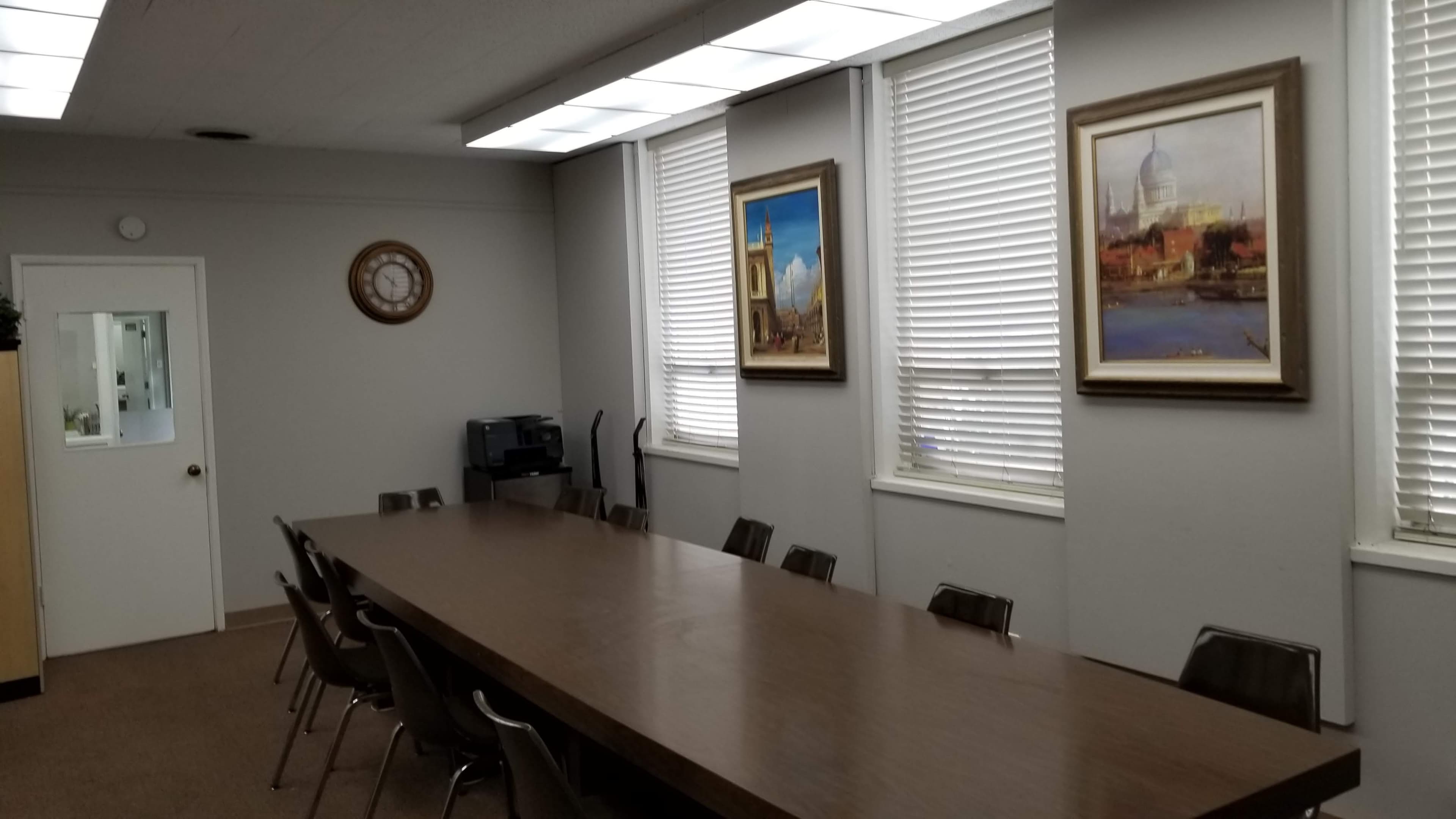 A long conference table with chairs is situated in a windowed meeting room adorned with two framed paintings.