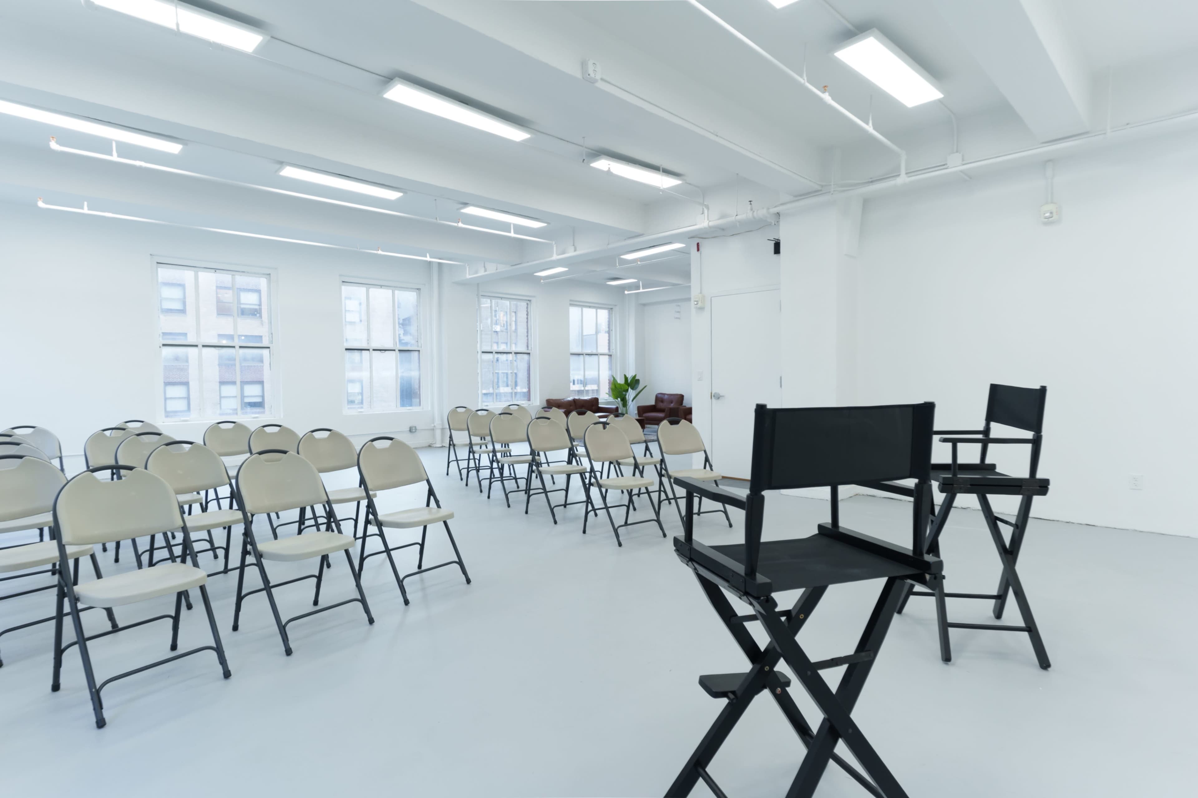 The image depicts an empty, brightly lit room with rows of beige folding chairs arranged in front of two director's chairs and a small table.