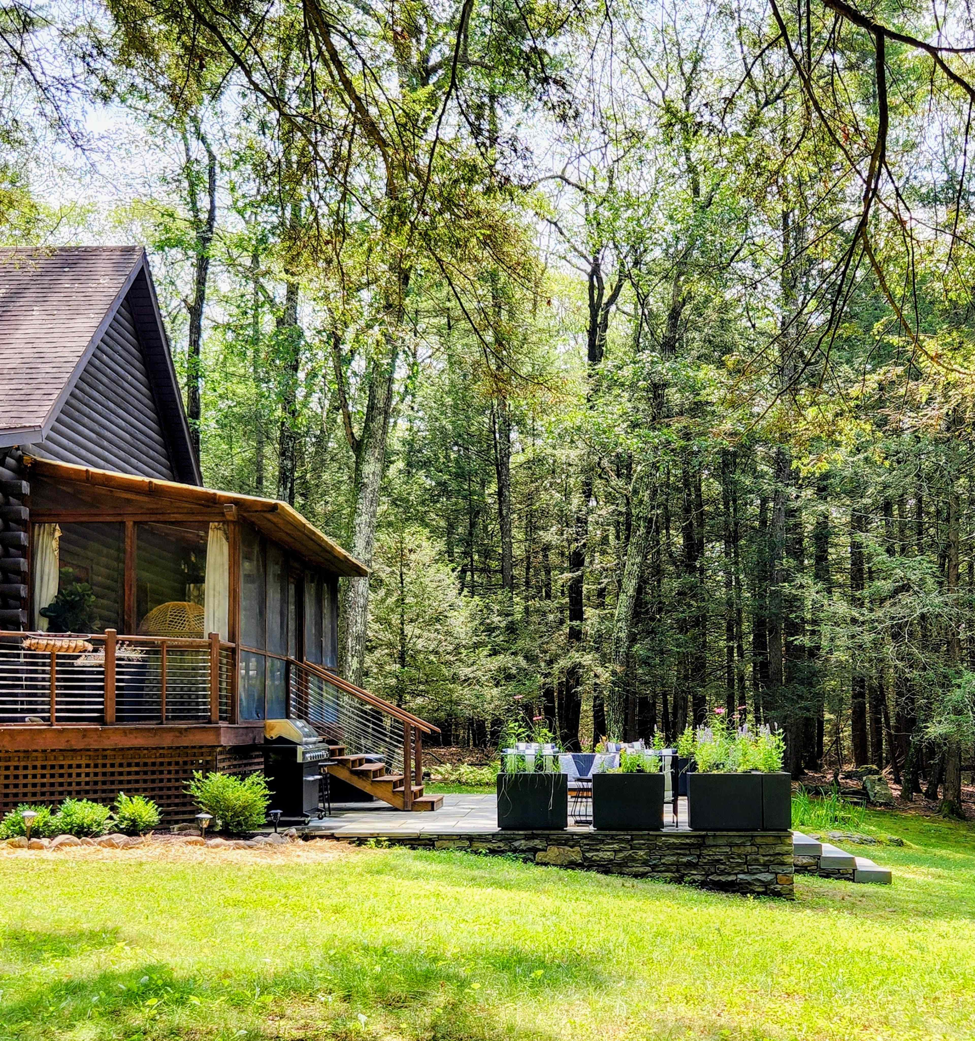 A wooden cabin is surrounded by dense trees, featuring a deck with seating and potted plants in the foreground.