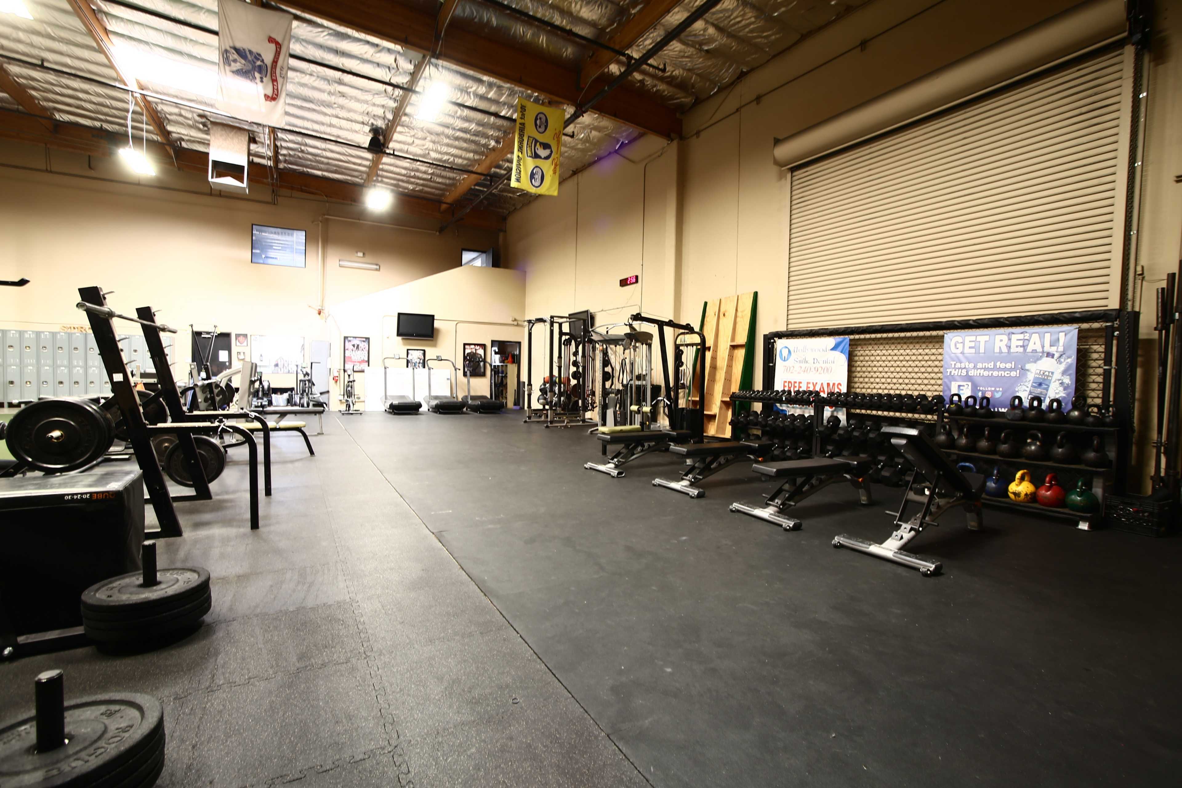 A spacious gym interior with various strength training equipment, including benches, dumbbells, and weightlifting platforms, along with lockers along one wall.