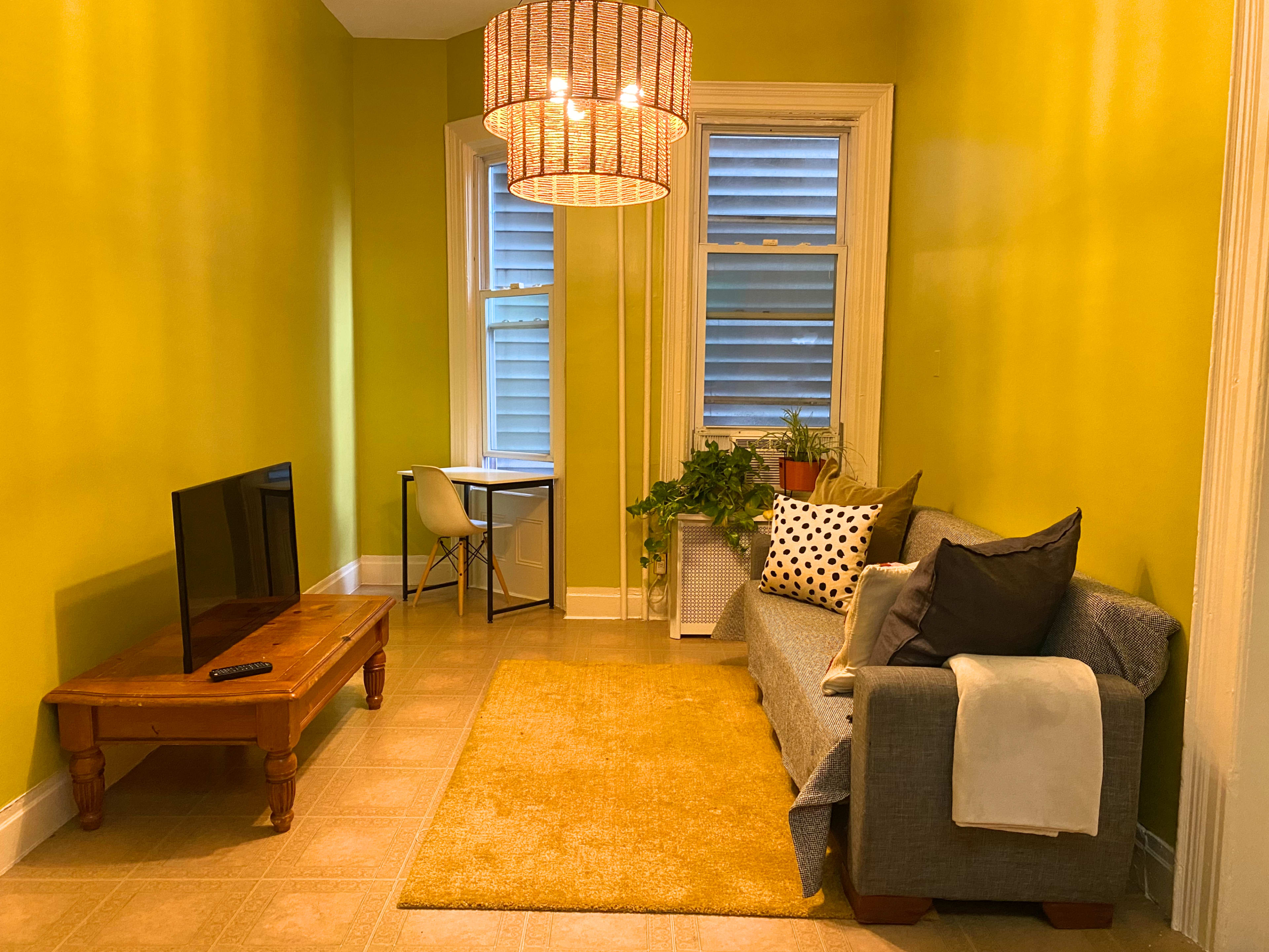 The image shows a small, brightly colored room with green walls, a sofa, coffee table, television, and a desk by the window.