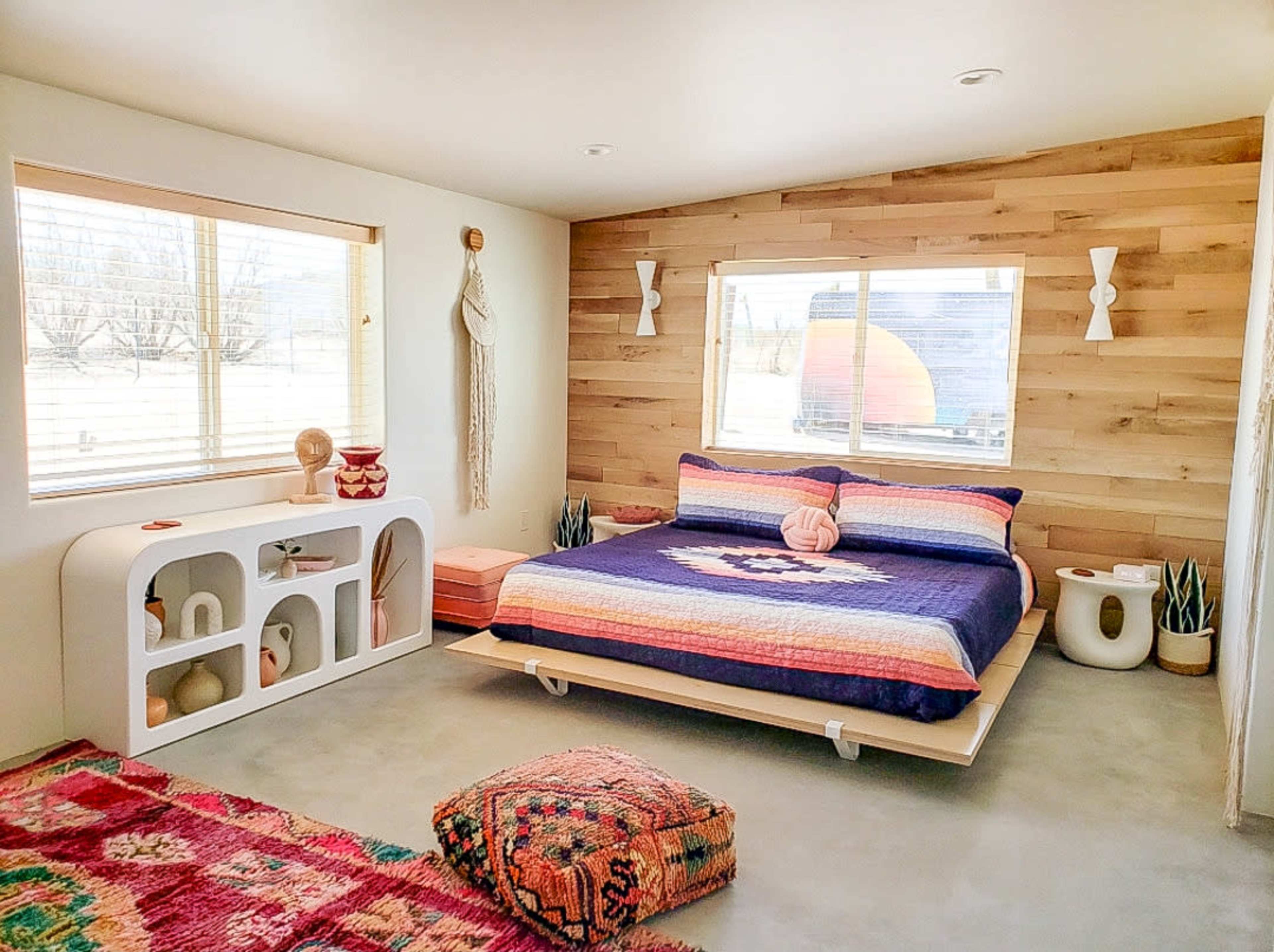 A bedroom features a bed with a colorful patterned blanket, modern furniture, and large windows allowing natural light to enter.