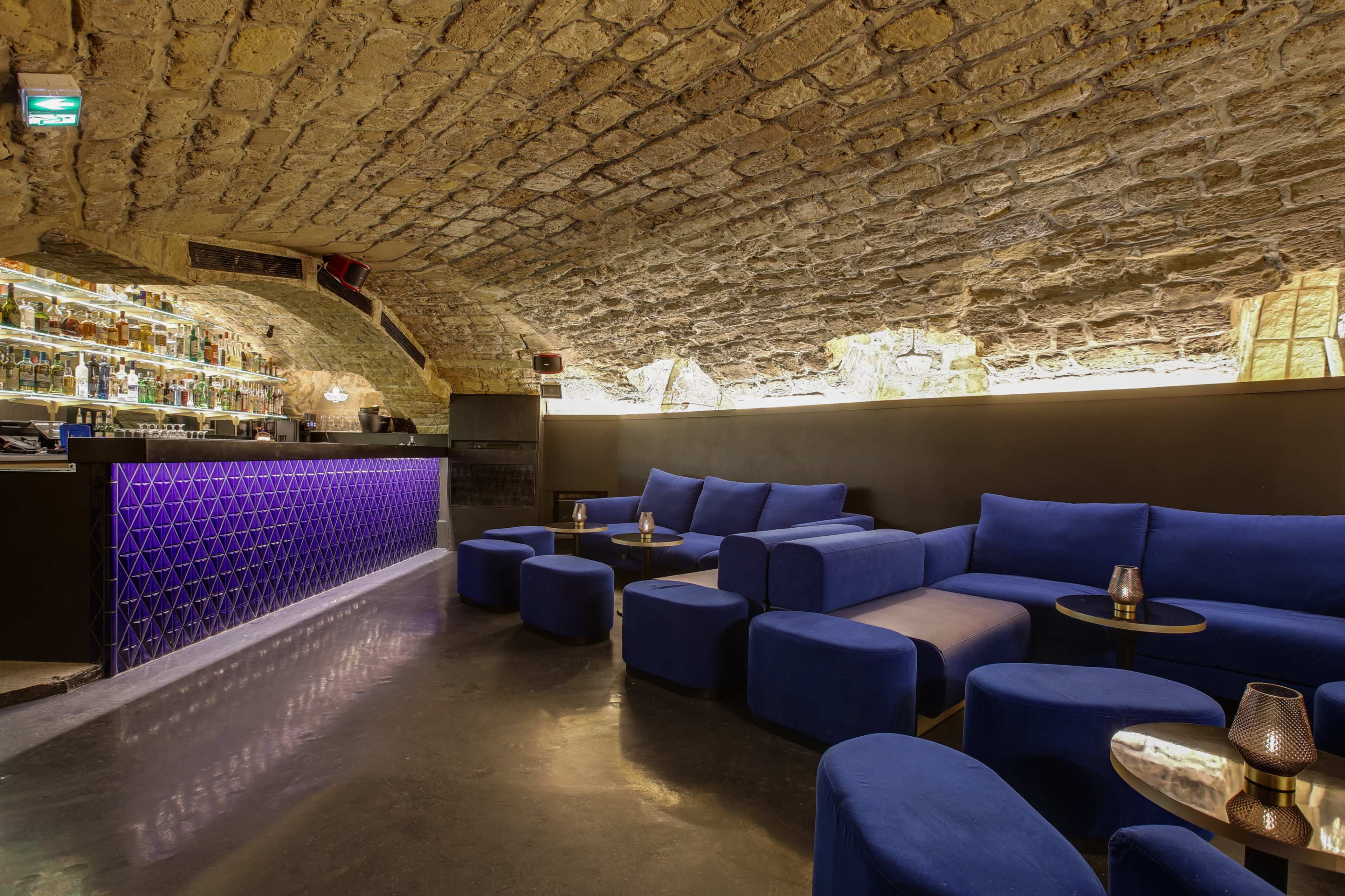 A dimly lit bar features a curved stone ceiling, a purple-lit backlit bar, and plush blue seating arrangements.