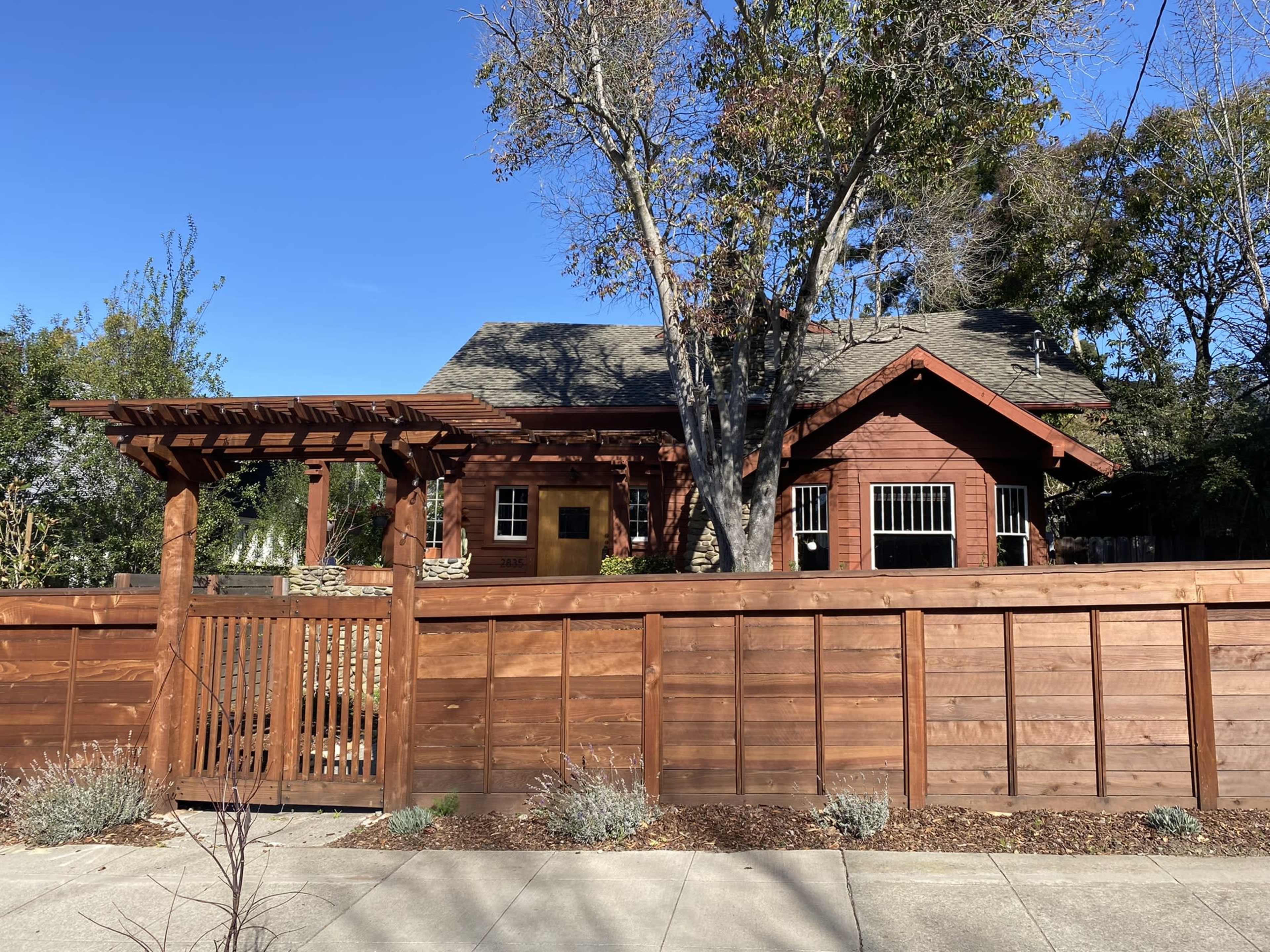 A wooden house with a pitched roof is surrounded by a low fence and features a pergola at the entrance.