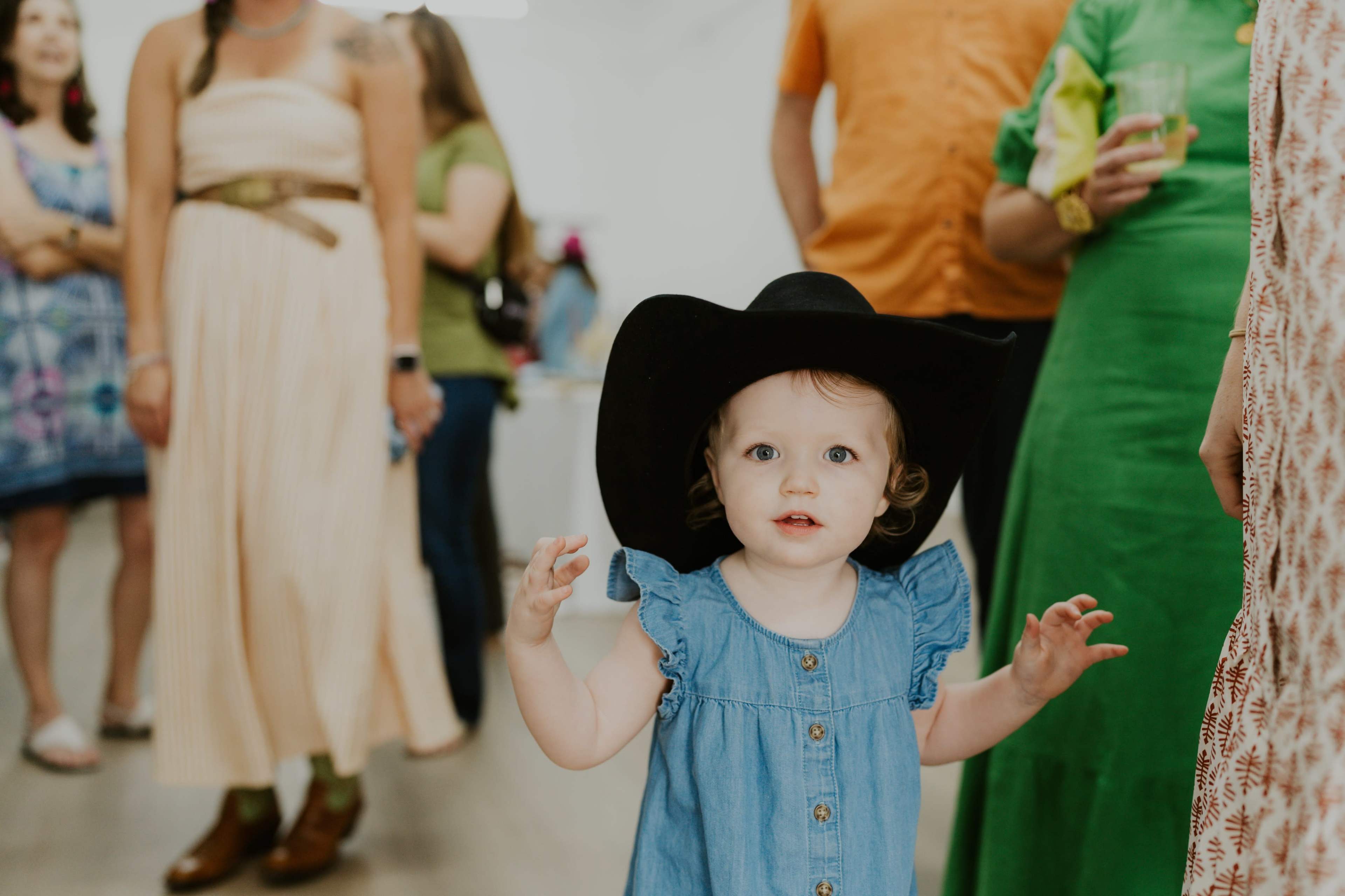A toddler wearing a large black cowboy hat stands in the foreground of an art gallery, while adults mingle in the background.