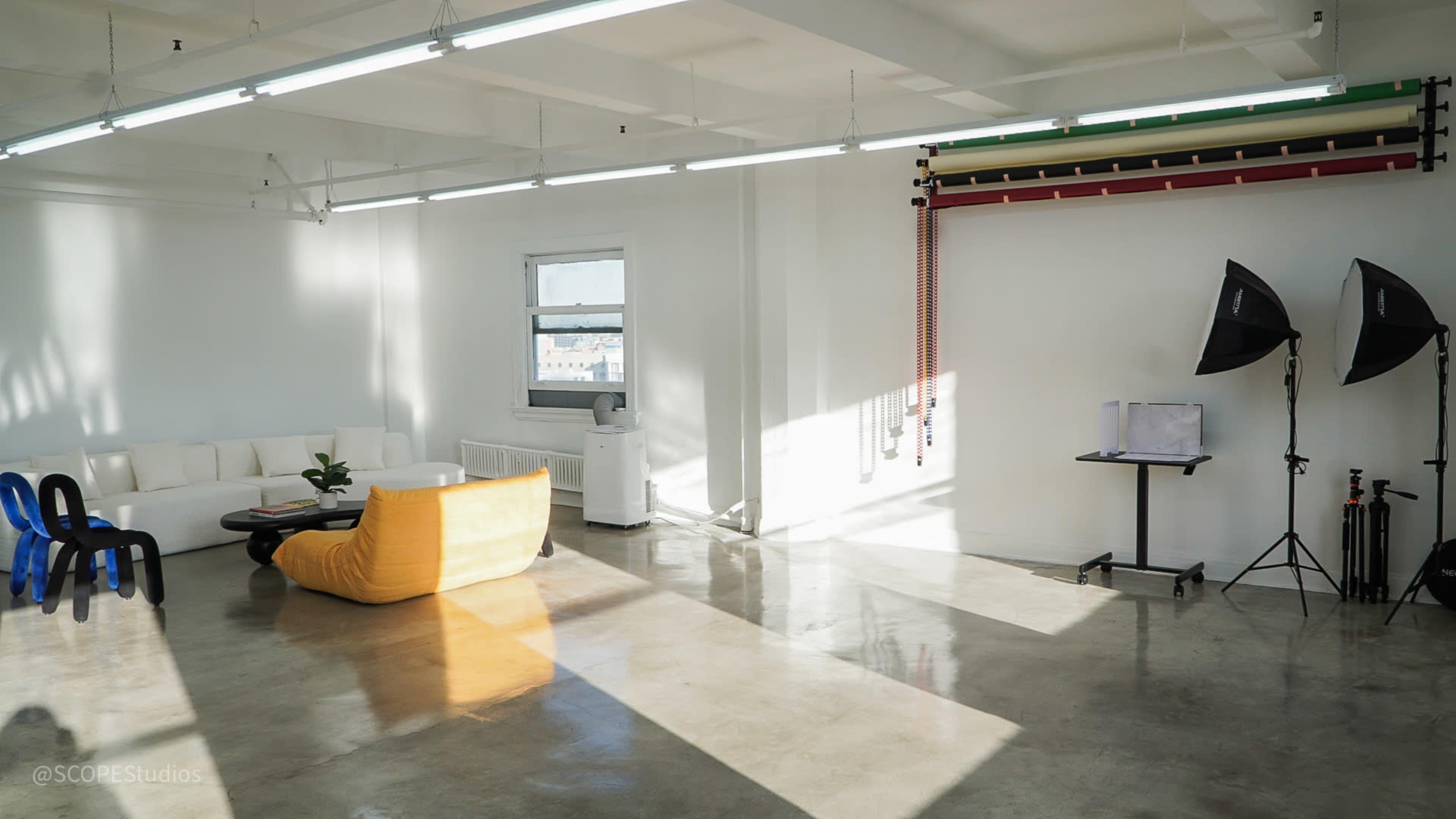 The image shows a bright, minimalist studio space with a large yellow sofa, a white couch, and studio lighting equipment set up against the wall.
