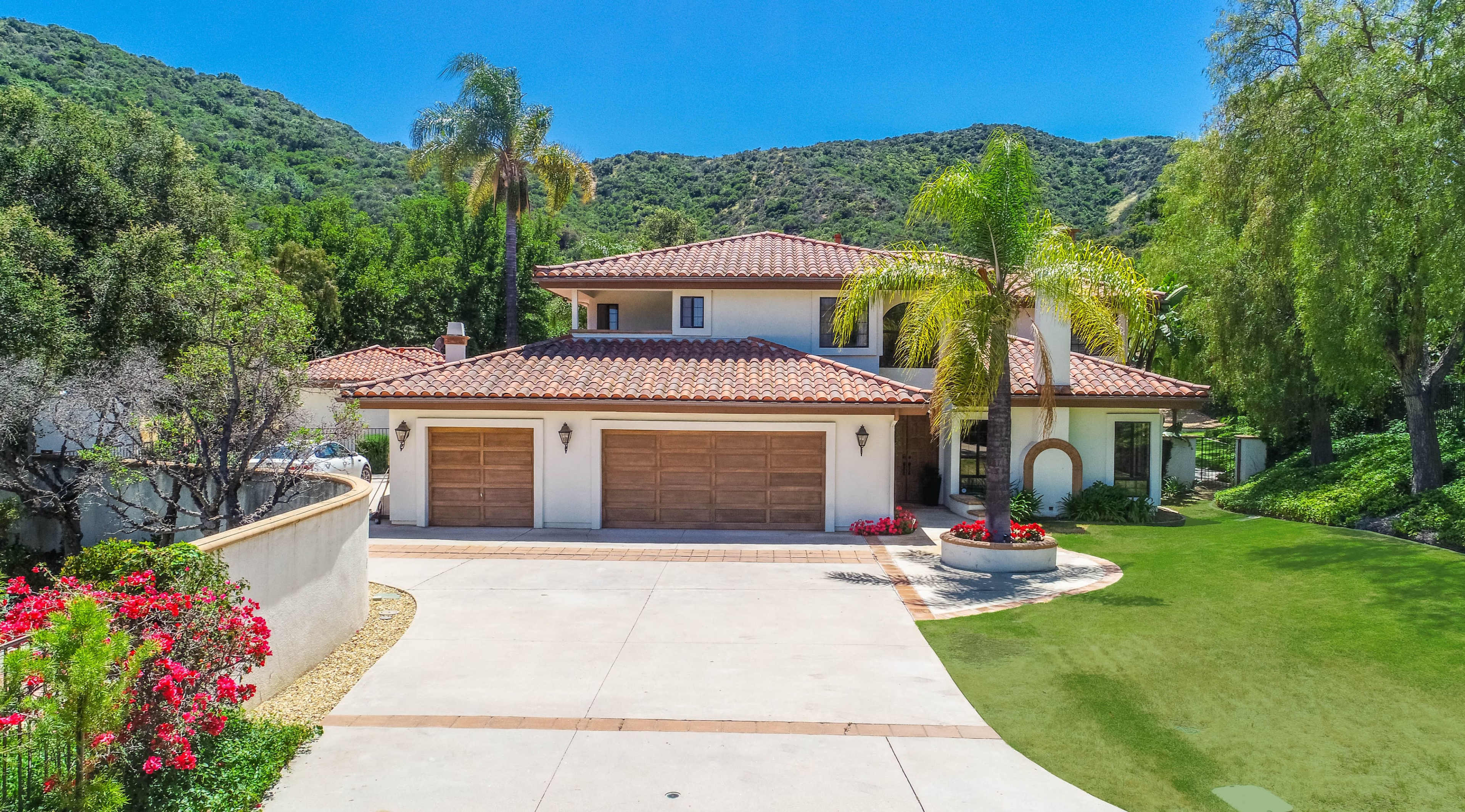 A two-story house with a tile roof is surrounded by greenery and features a paved driveway with two wooden garage doors.