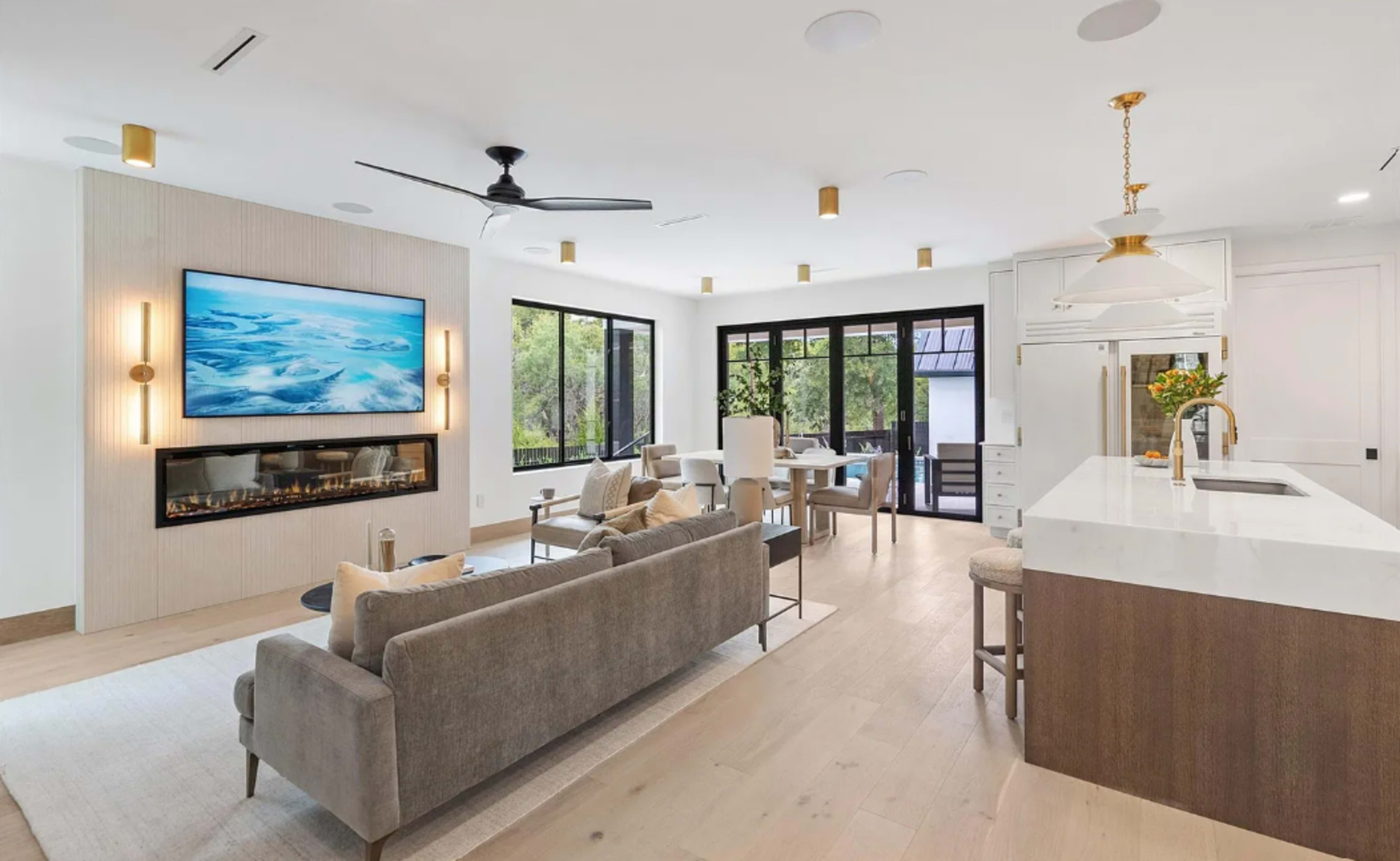 The image shows a modern open-concept living space featuring a living area with a couch and television, a dining area with a table, and a kitchen with a large island.