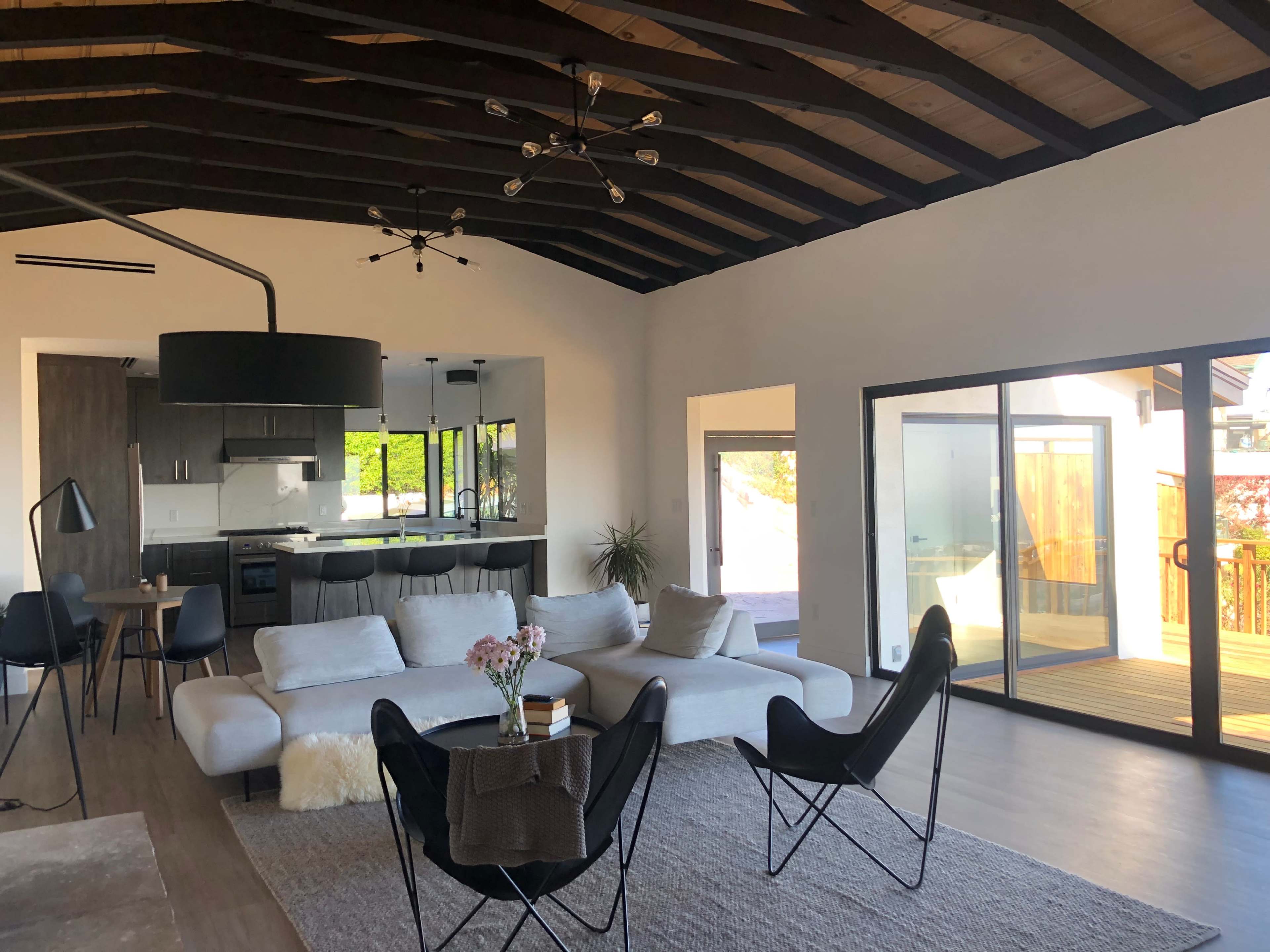 A modern living room with a large sofa, two black chairs, a dining table, and a kitchen area in the background, all featuring high ceilings with exposed beams and large windows.