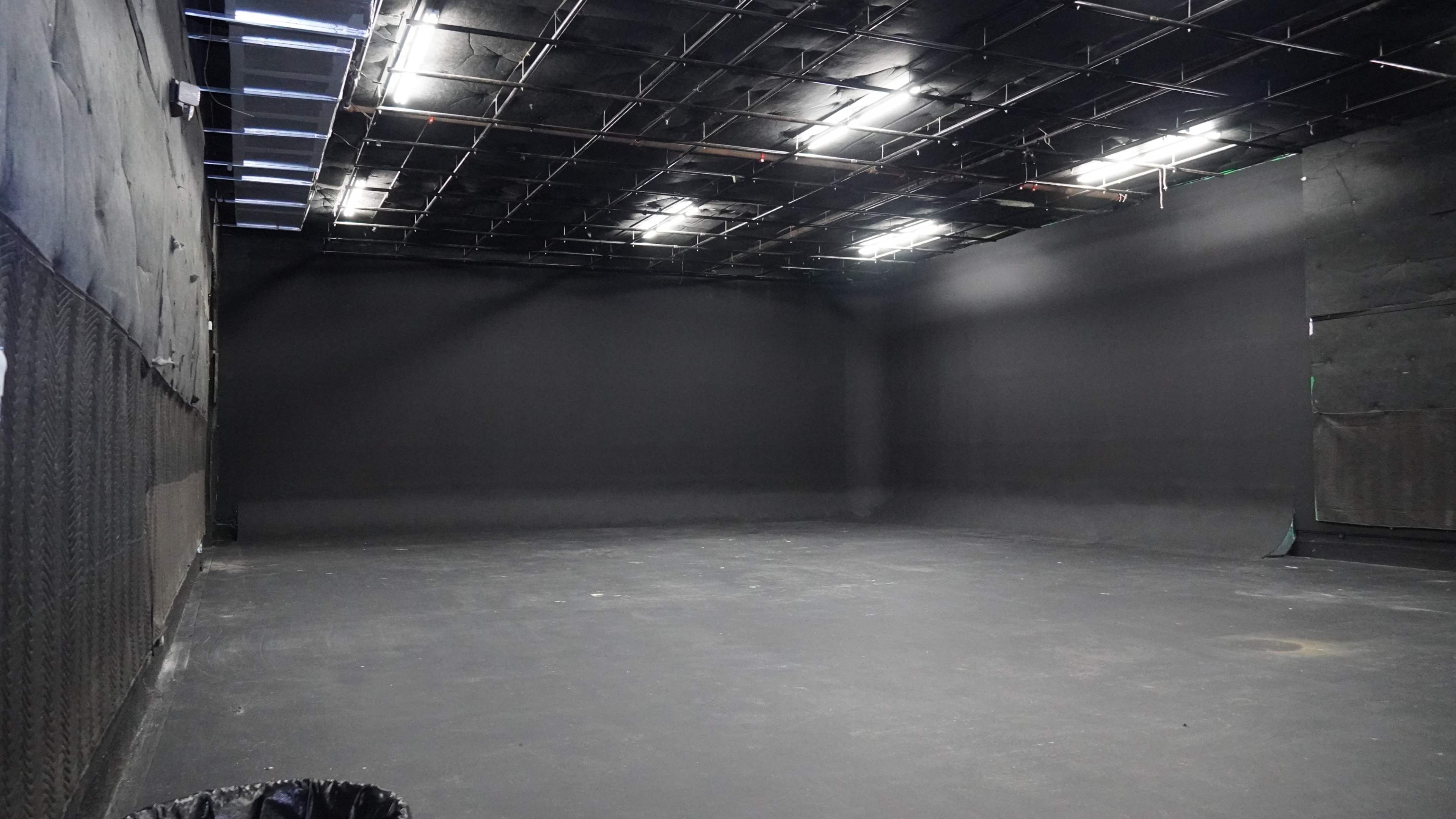 The image shows an empty studio space with black walls and a dark floor, illuminated by overhead lights.
