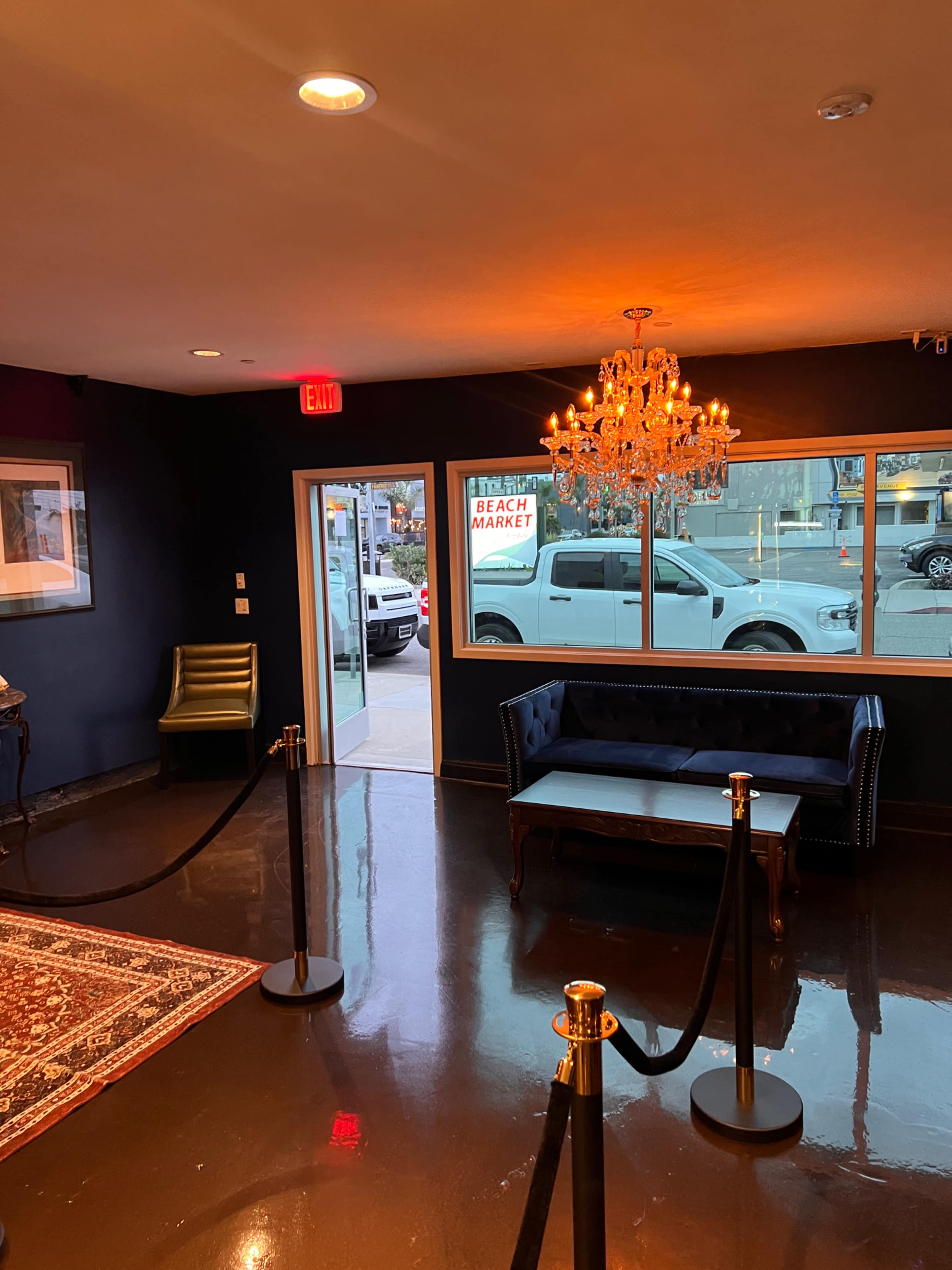 The interior of a lobby features a blue velvet couch, a chandelier, and velvet ropes lining the entrance, with a view of a street and a market beyond the windows.