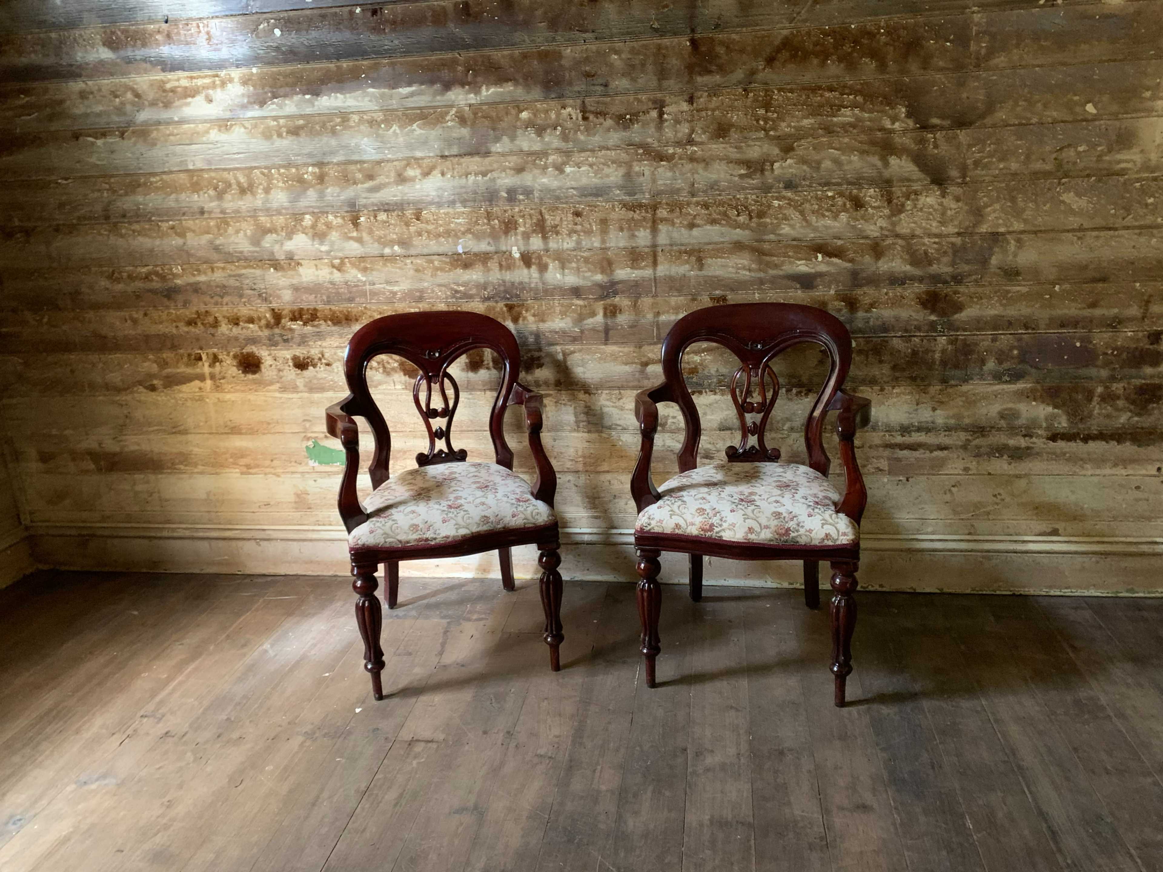 Two wooden chairs with floral upholstery are positioned against a weathered wooden wall in a dimly lit room.