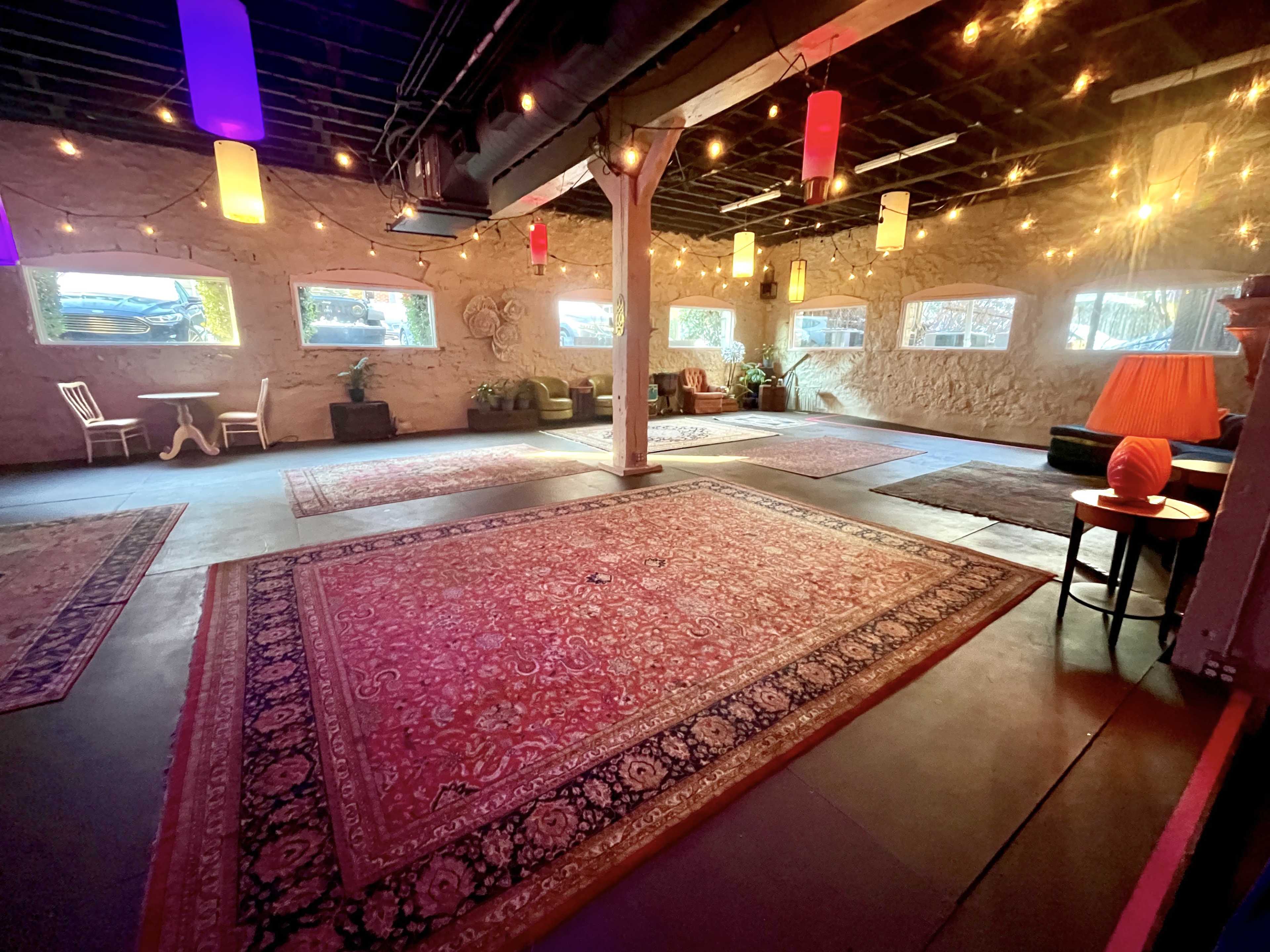 The image shows a spacious, softly lit interior with area rugs on the floor, string lights hanging from the ceiling, and colorful lamps illuminating various seating areas.