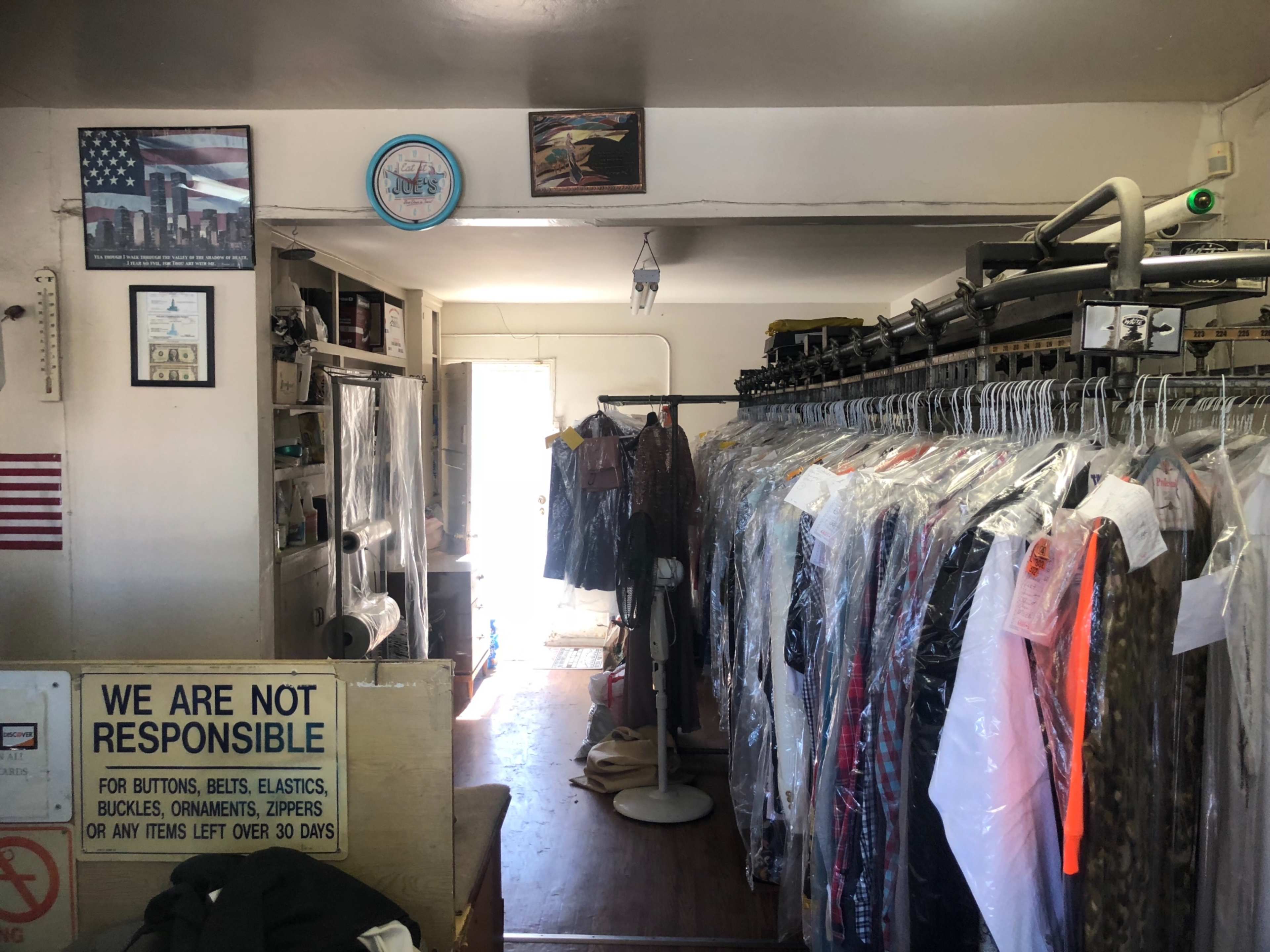 The interior of a dry cleaning shop featuring clothing on hangers, shelving units, and a sign stating "WE ARE NOT RESPONSIBLE" near the entrance.