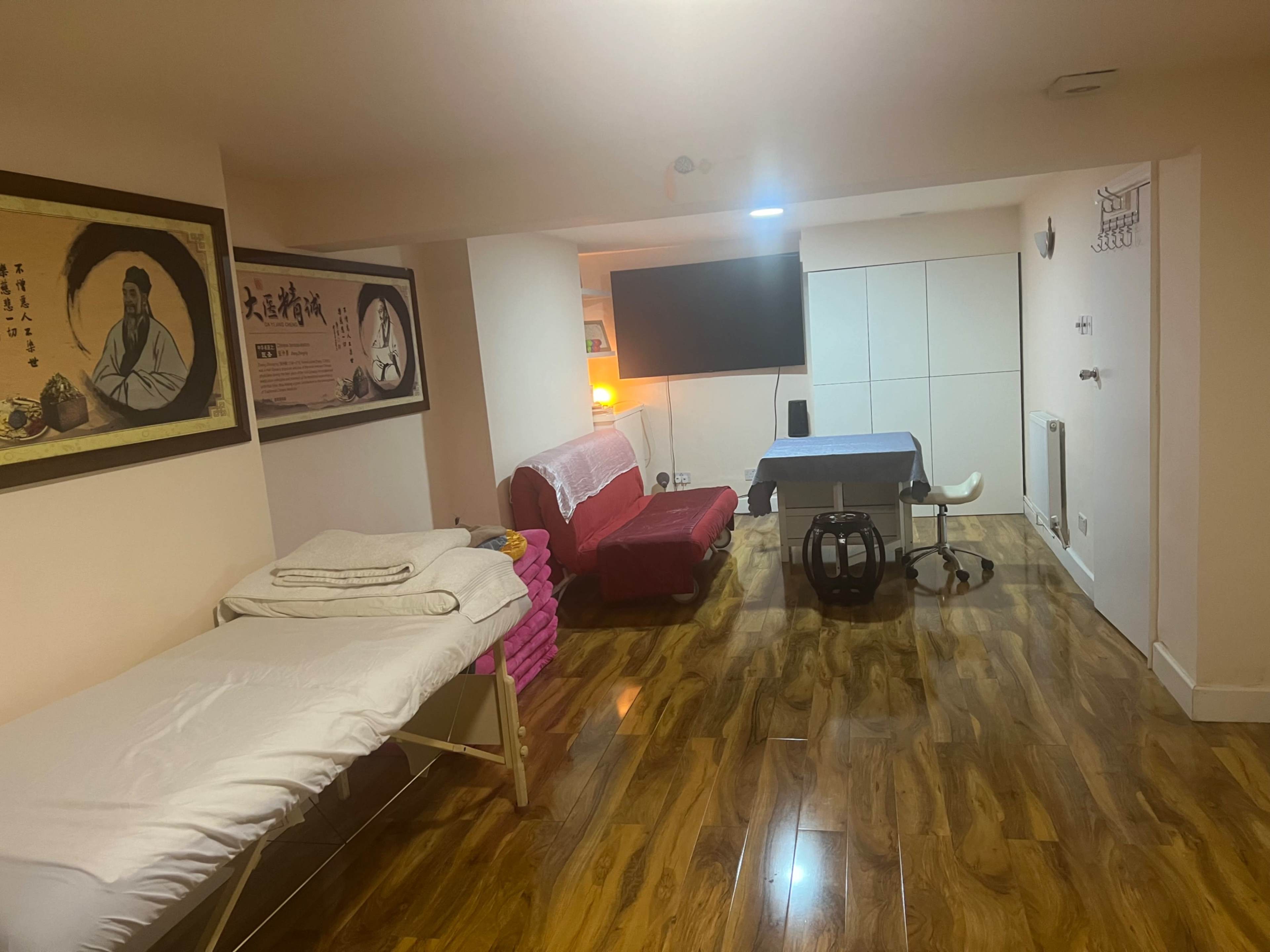 The image shows a therapy or treatment room with a massage table, a desk with a chair, a red couch, and a large wall-mounted screen.